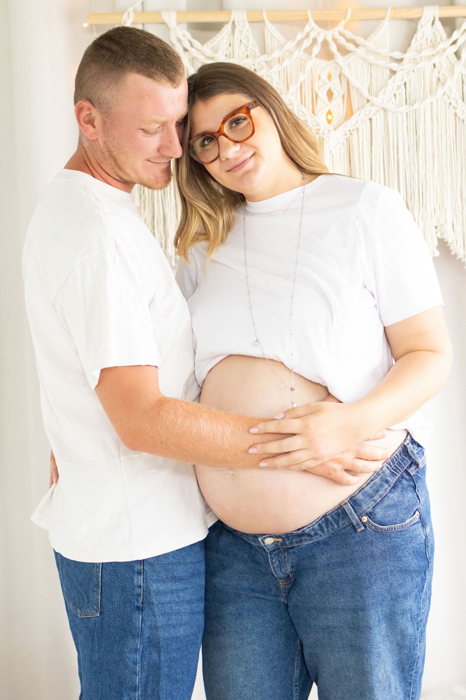 Couple in white tees and jeans posing for maternity photo, man embracing pregnant woman from behind indoors.