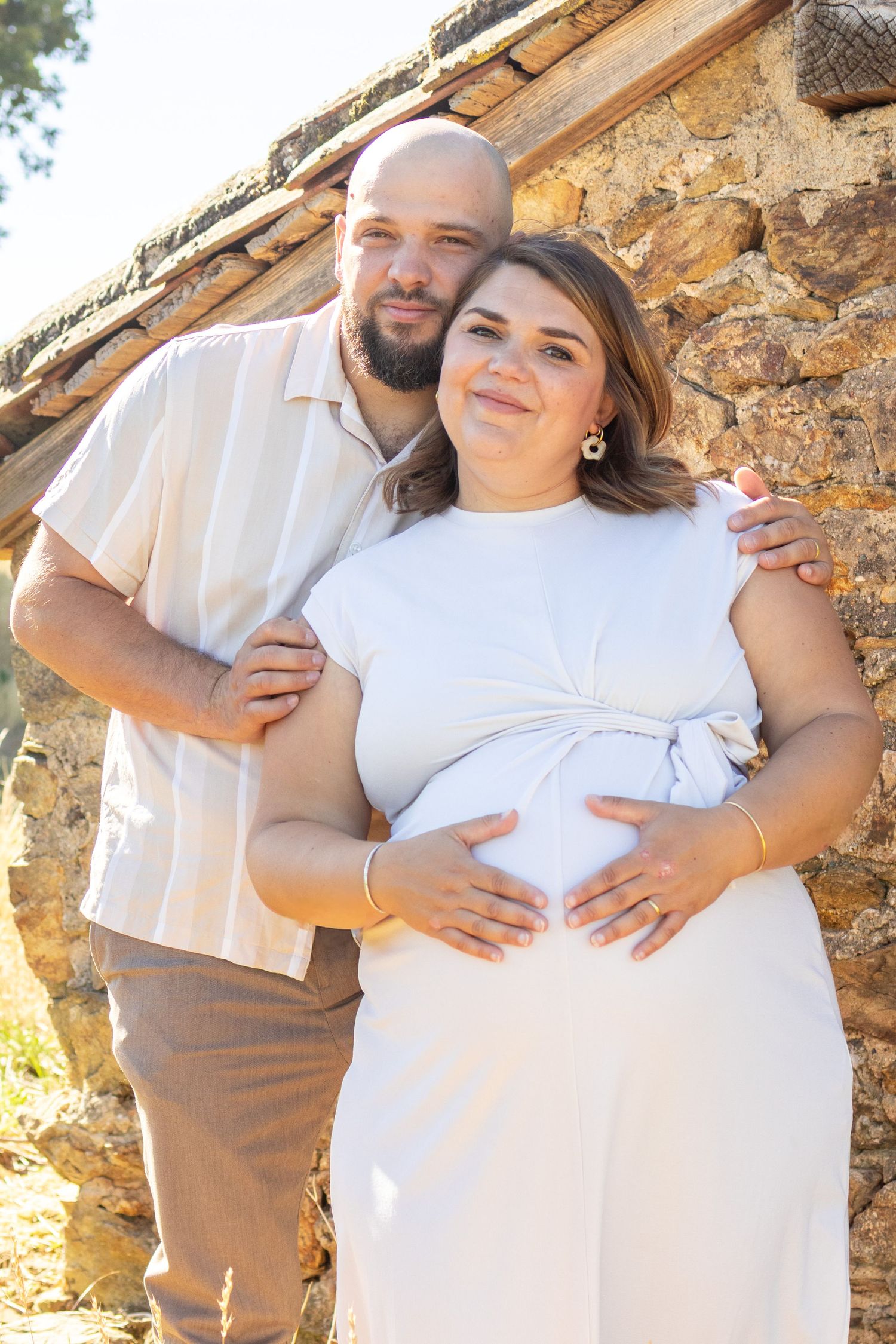 A bald man embraces a pregnant woman in a white dress against a rustic stone wall outdoors.