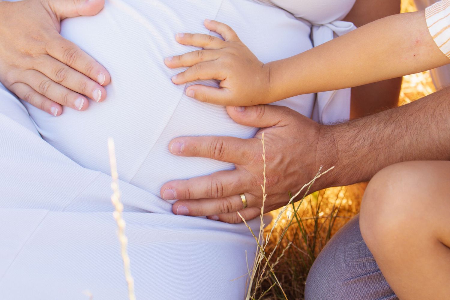Family hands resting on pregnant woman's belly outdoors in warm sunlight, symbolizing love and new life.