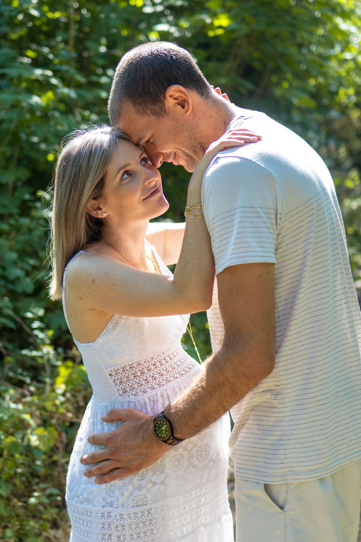 A loving couple embraces outdoors, the woman in a white lace dress smiling up at the man against a lush green background.