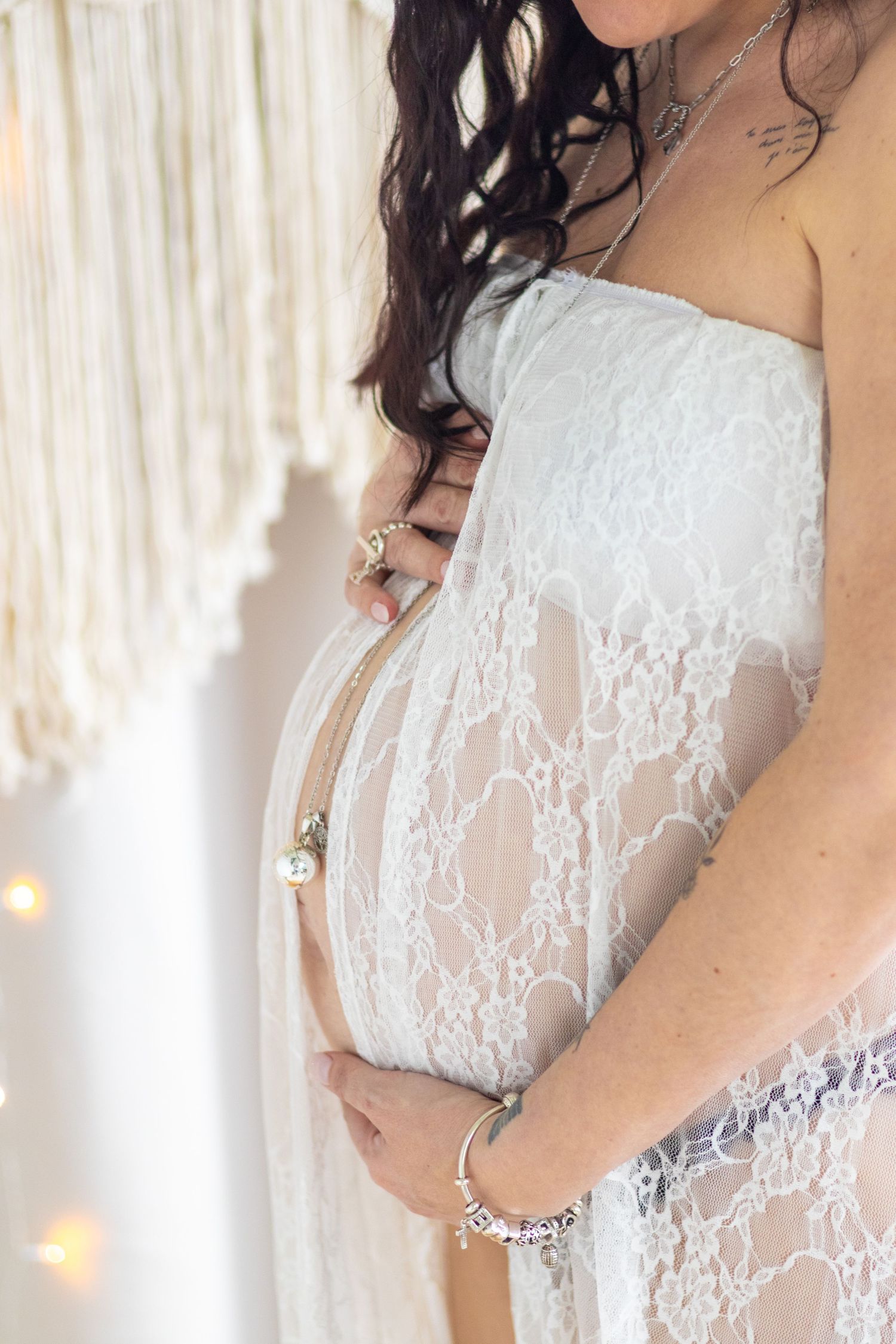 Pregnant woman in white lace maternity gown cradling her baby bump, boho decor backdrop.