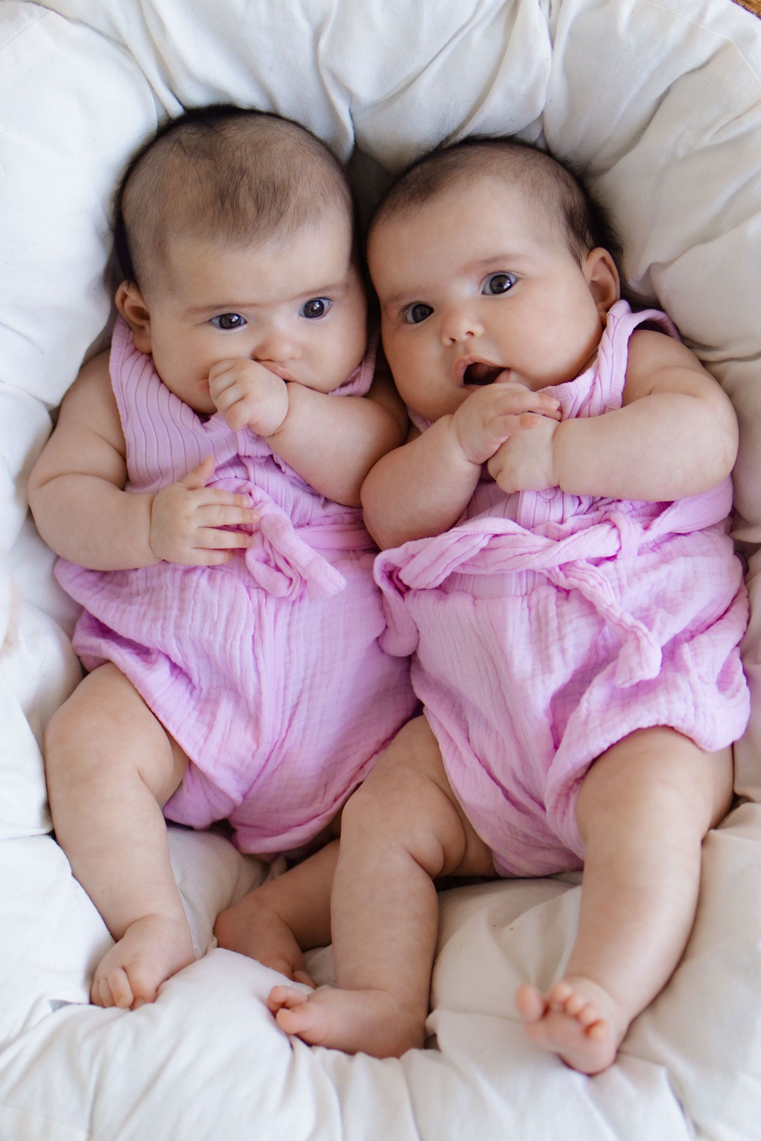 Two adorable twin baby girls in matching pink muslin rompers lying together on a white fluffy surface.
