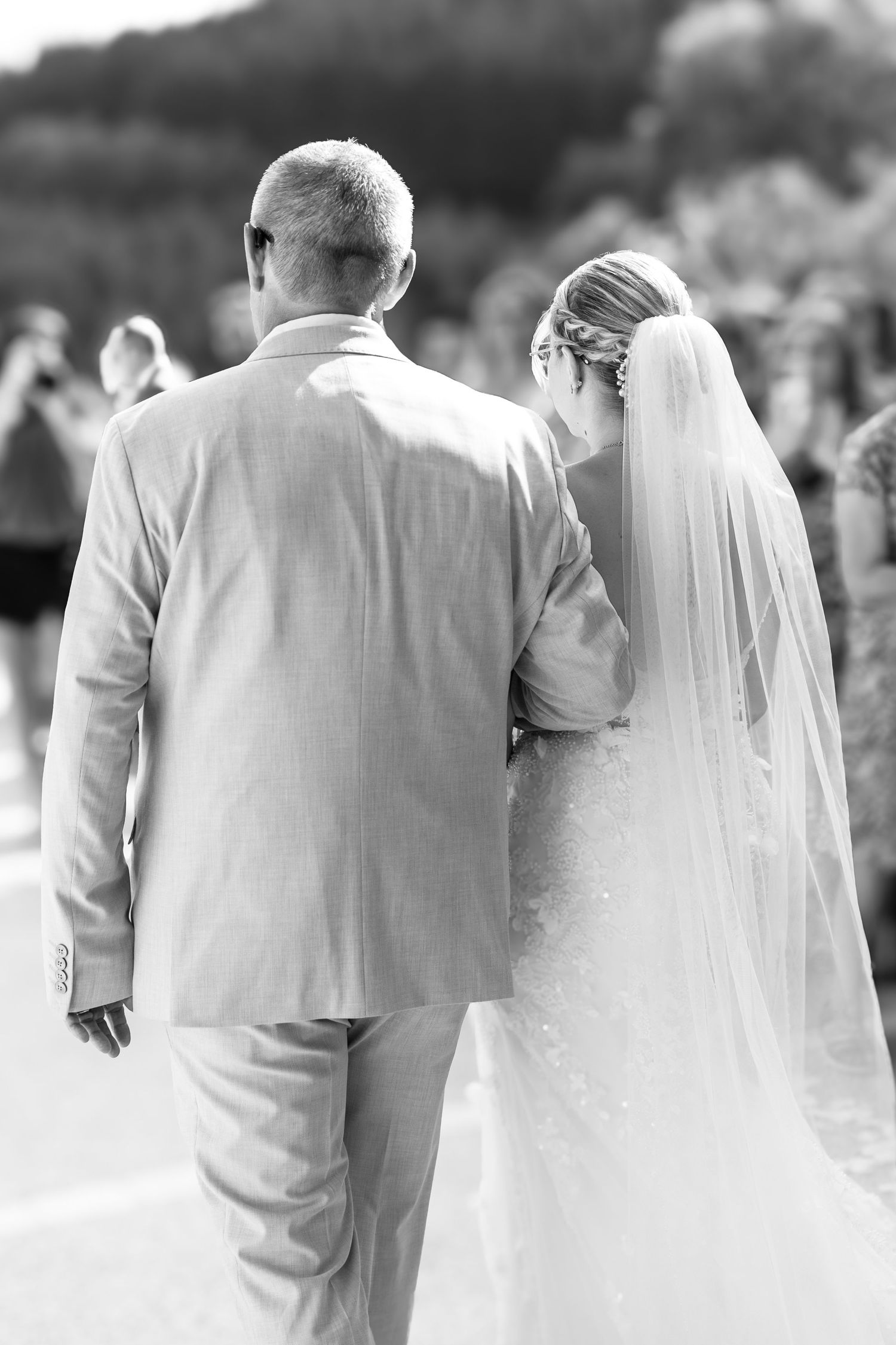 Black and white photo of a bride in veil and groom in suit walking together from behind at outdoor wedding.