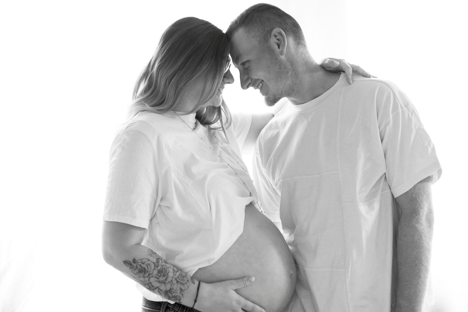 Black and white maternity photo of a pregnant couple in white t-shirts tenderly touching foreheads while cradling baby bump.