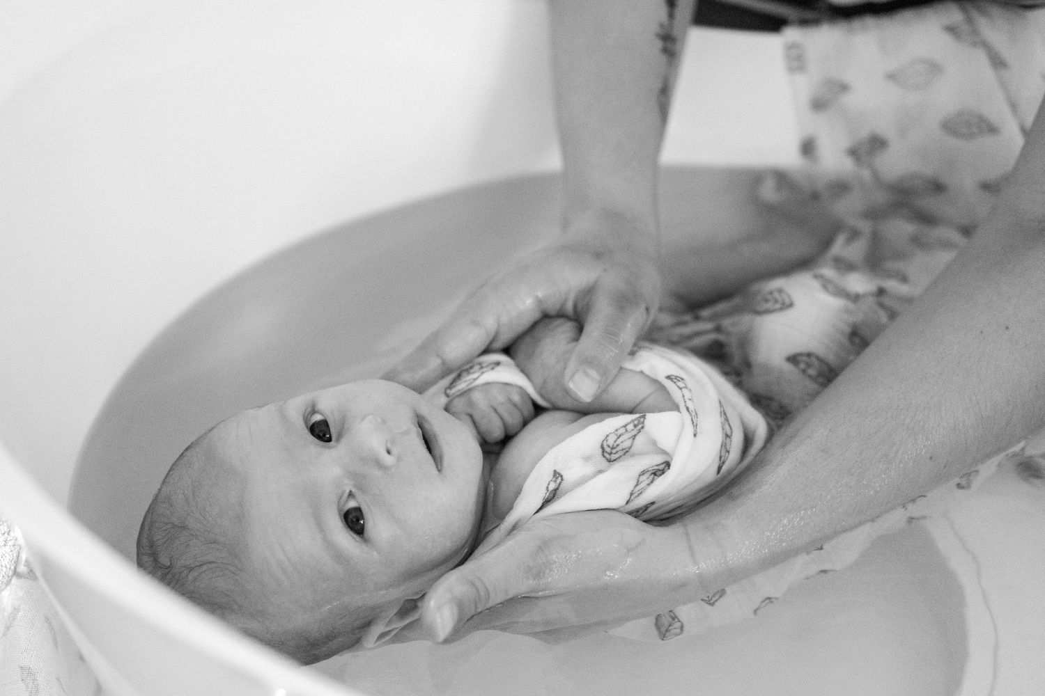 Black and white photo of a newborn baby being bathed in a white infant tub by caring adult hands.