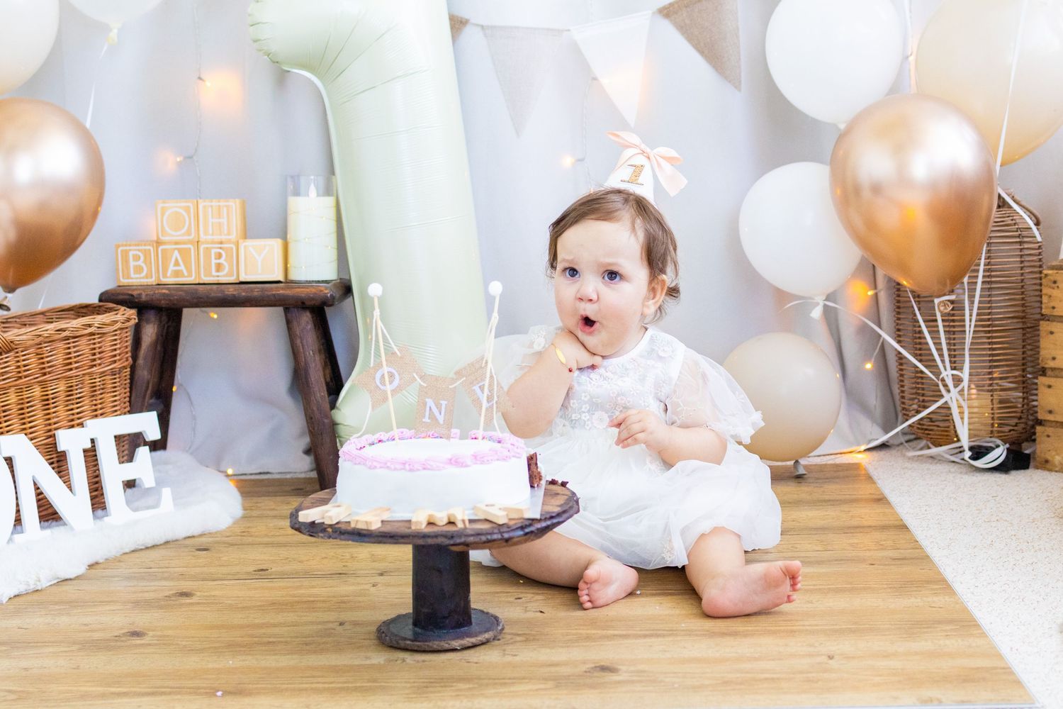 Baby girl in white dress sits beside a pink first birthday cake, surrounded by balloons and ONE decorations.
