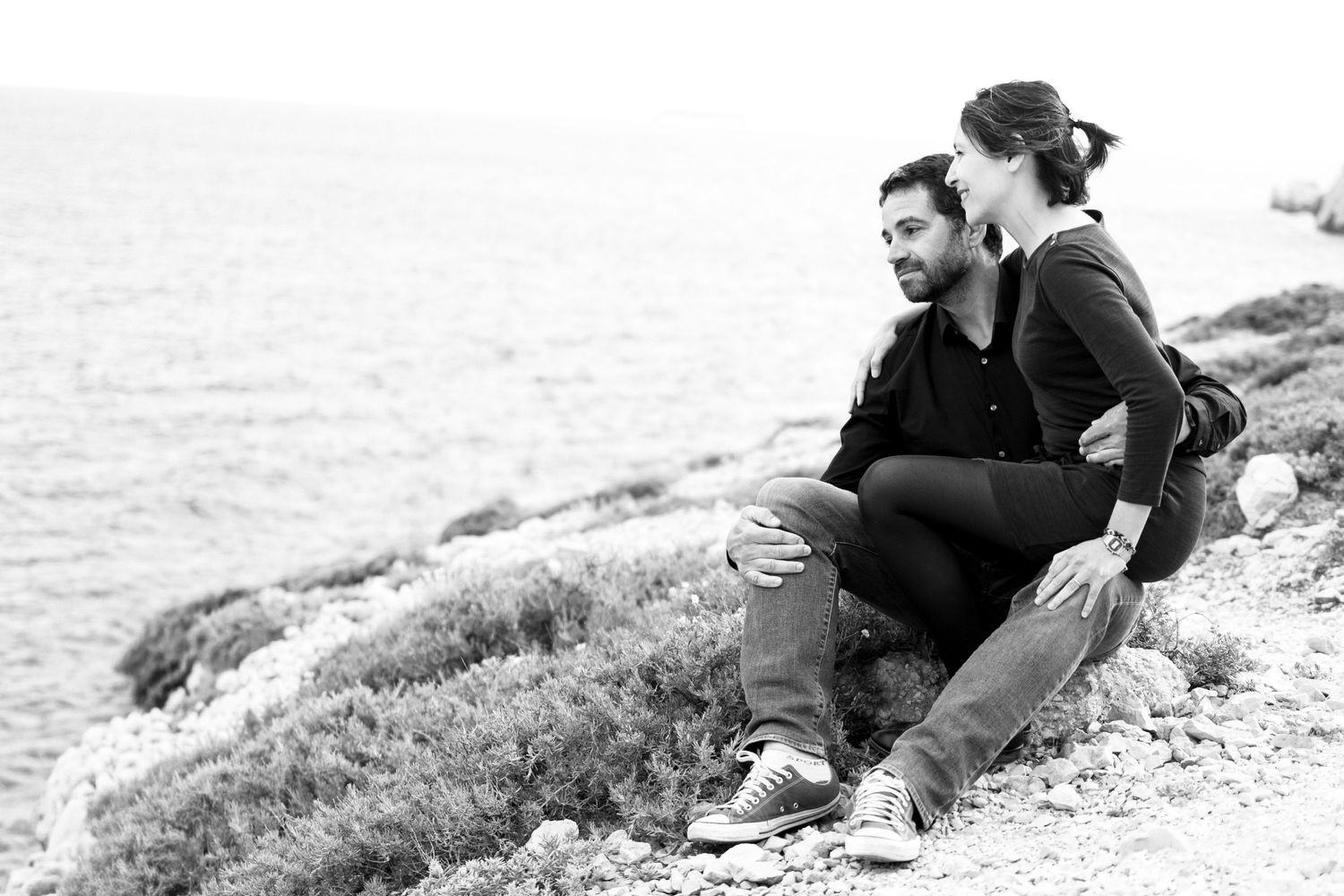 A couple sits together on a rocky coastal hillside, gazing at the sea in a black and white photo.