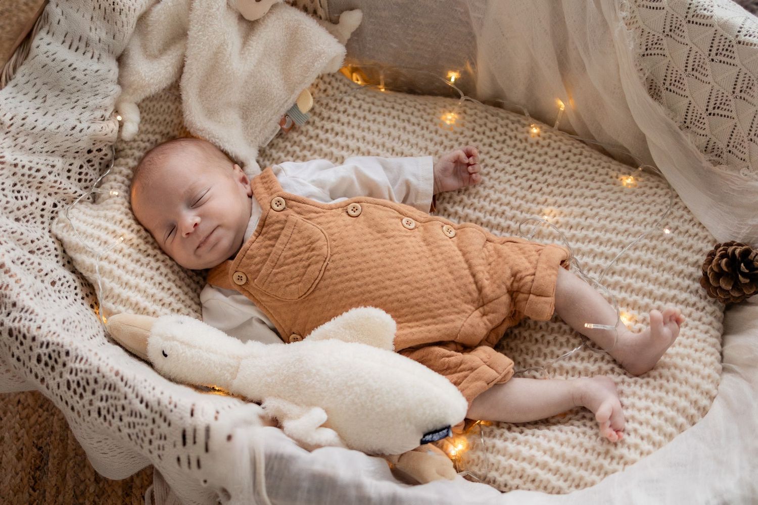 Newborn baby in orange knit romper lying in cozy basket with stuffed bunny toy and warm fairy lights.
