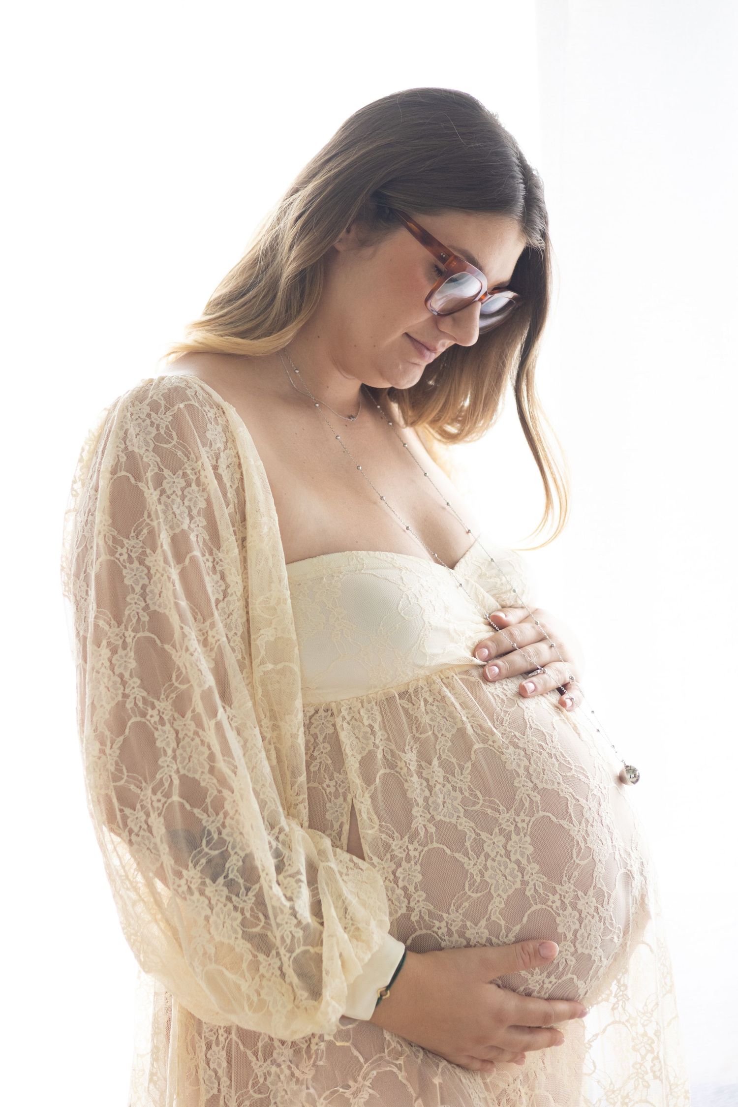 Pregnant woman in cream lace robe cradling baby bump, wearing glasses, soft white background, maternity photo.