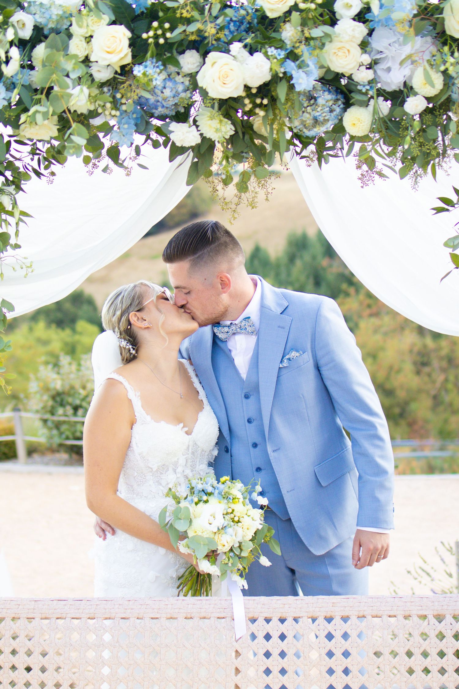 Bride and groom share first kiss under floral arch with blue hydrangeas and white roses at outdoor wedding ceremony.