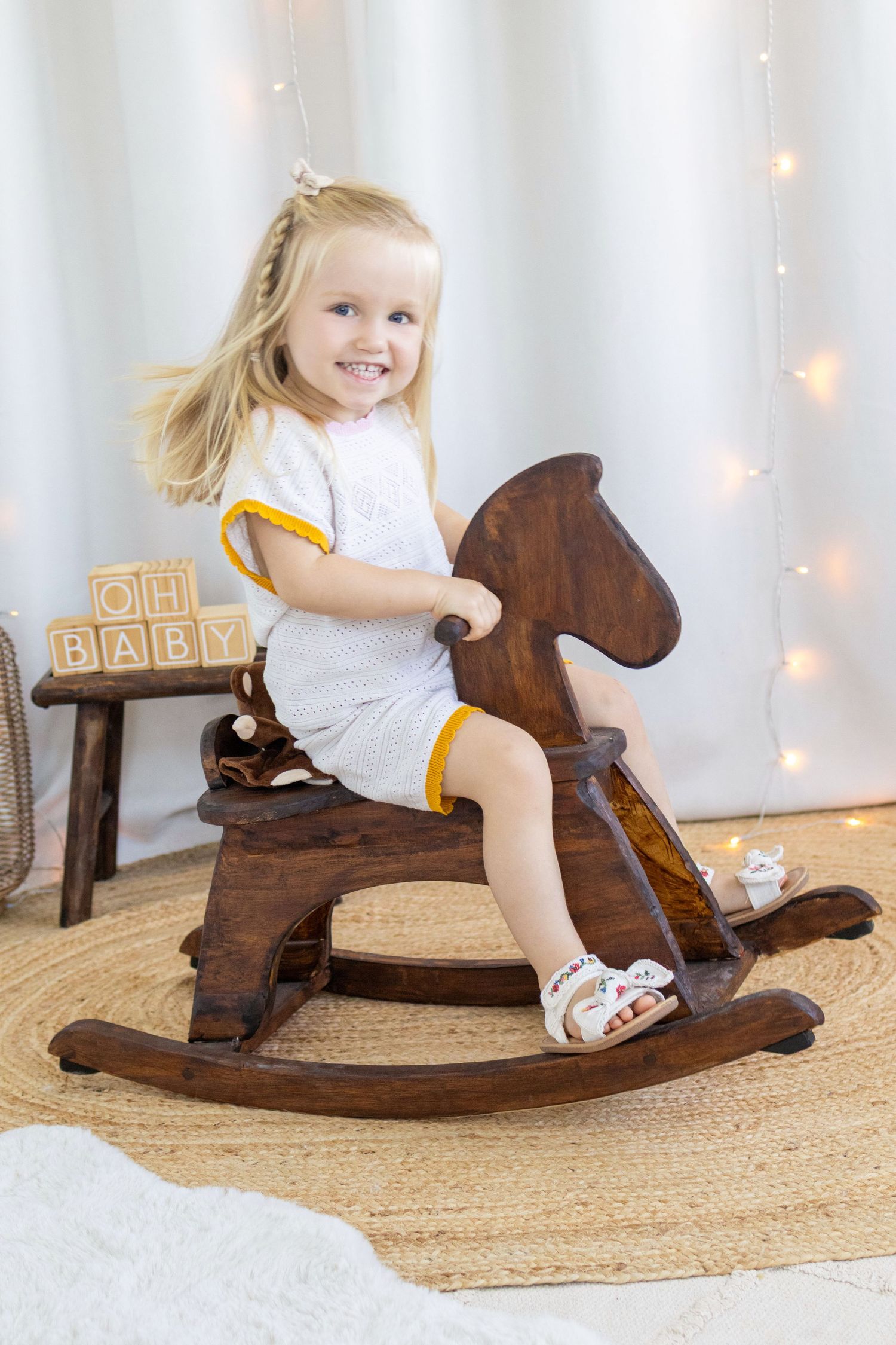 Smiling toddler girl with blonde hair riding a wooden rocking horse in a bright nursery room with fairy lights.