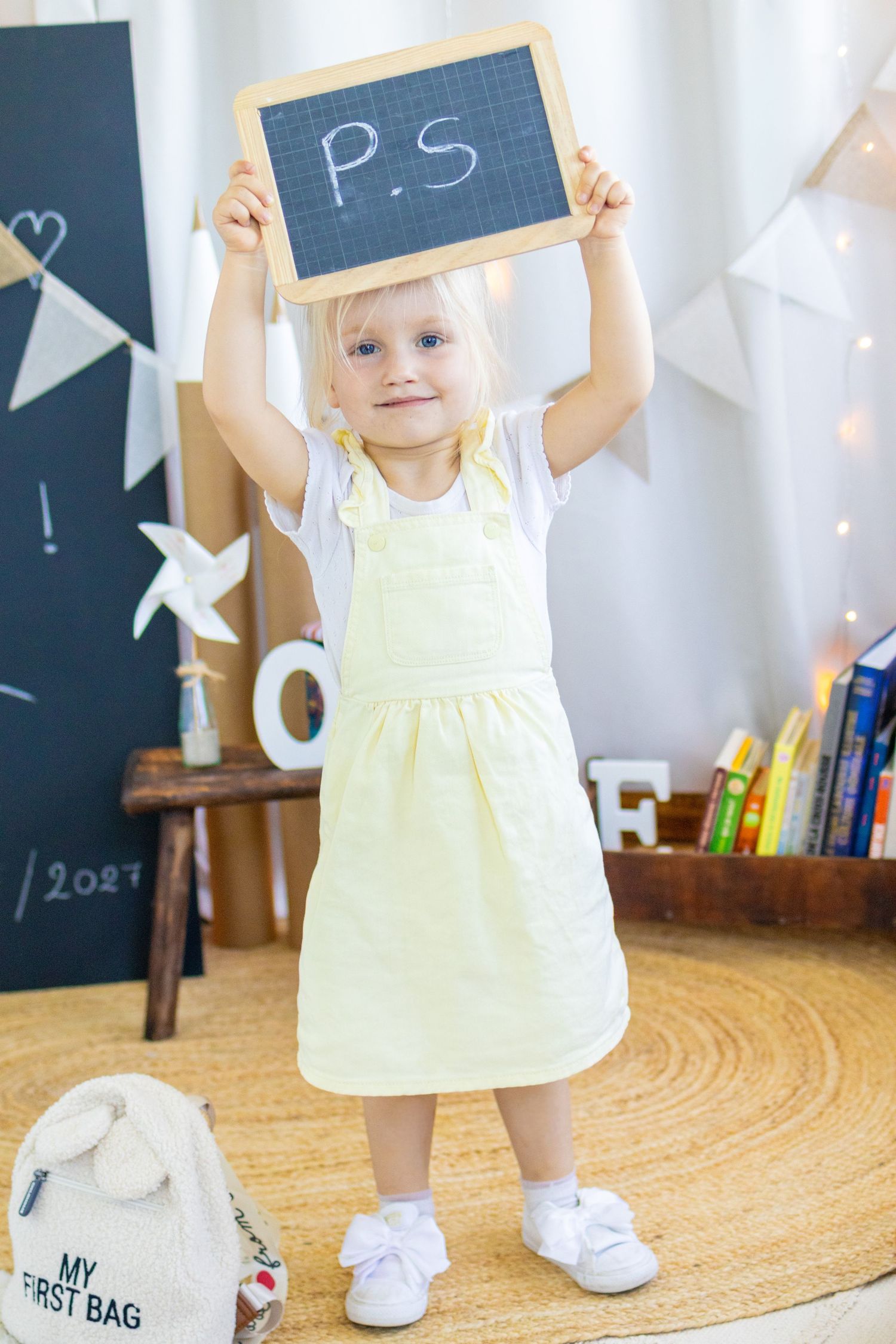 Cute blonde toddler girl in yellow dress holding a small chalkboard with P.S. written on it above her head.