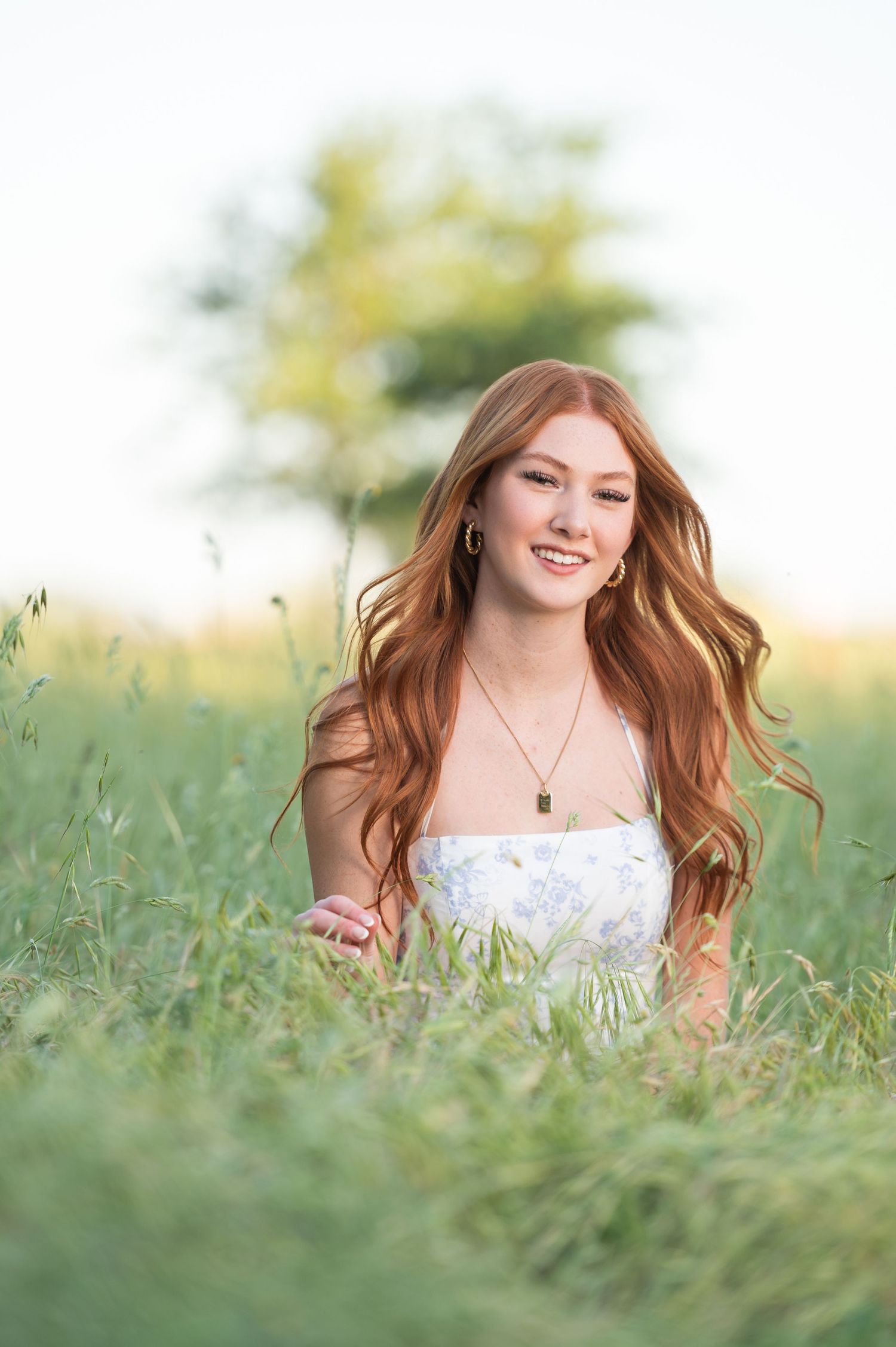 High School senior girl sitting in tall green grass with the wind blowing her red hair a bit.