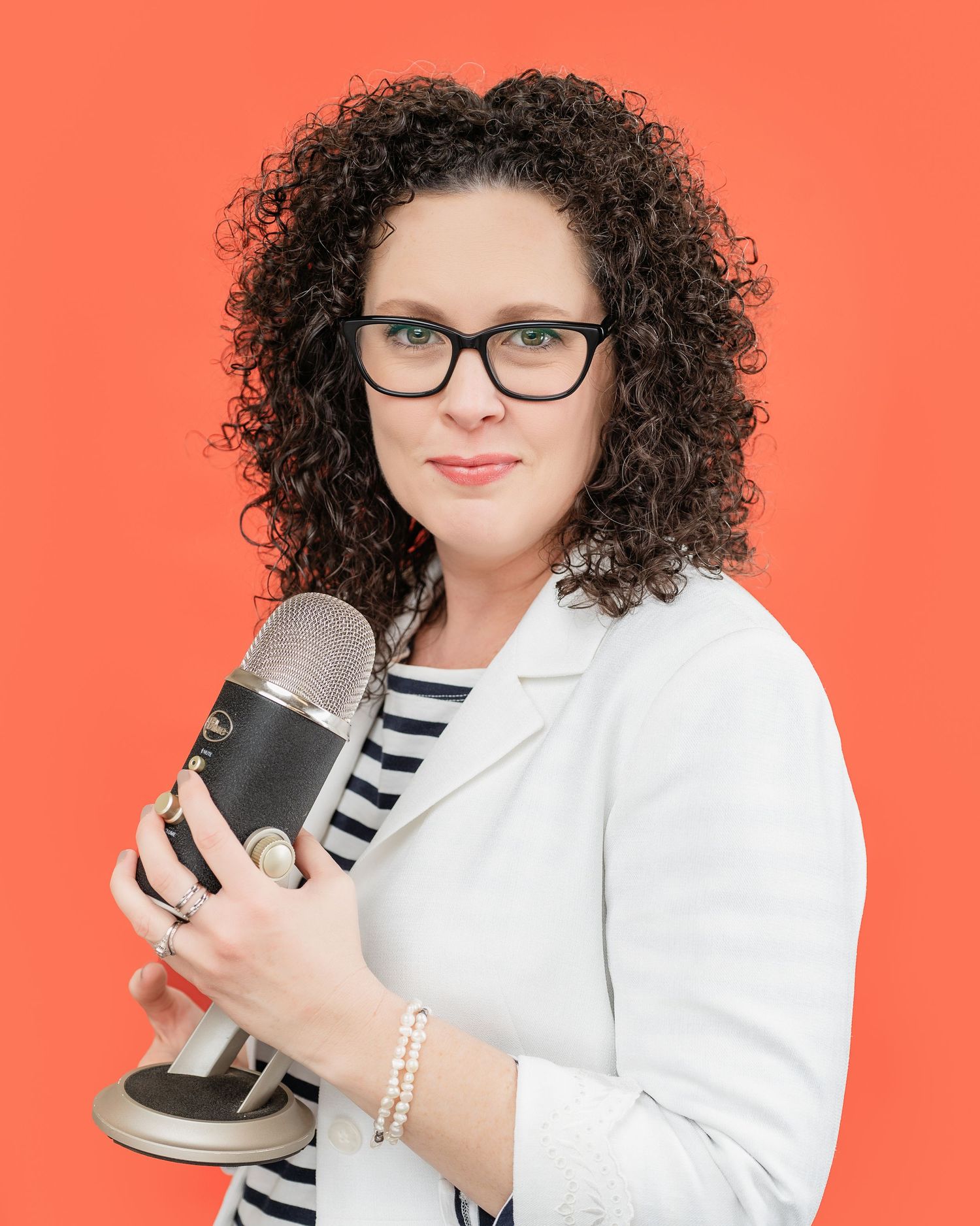 Headshot of a woman wearing black and white, holding a large microphone with a bright orange background
