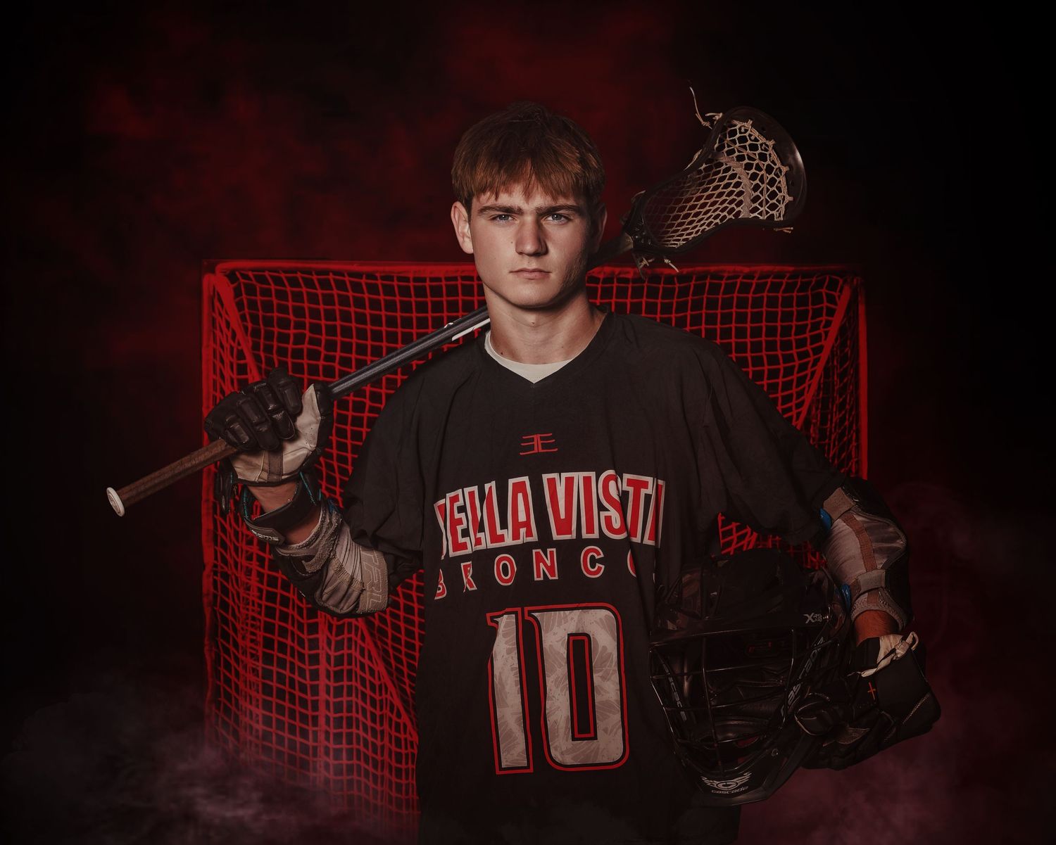 A Sportrait.  A grungy photo of a boy in a lacrosse uniform holding his stick, in front of the net.