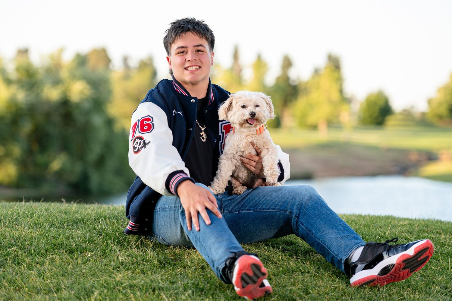Senior Boy wearing a letterman jacket, sitting on a grassy hill with pond in the background, while holding his small white dog