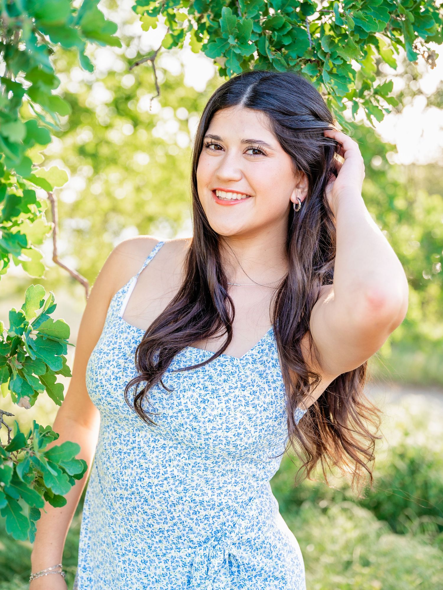 A senior girl photo with her hand in her hair, taken among green trees.