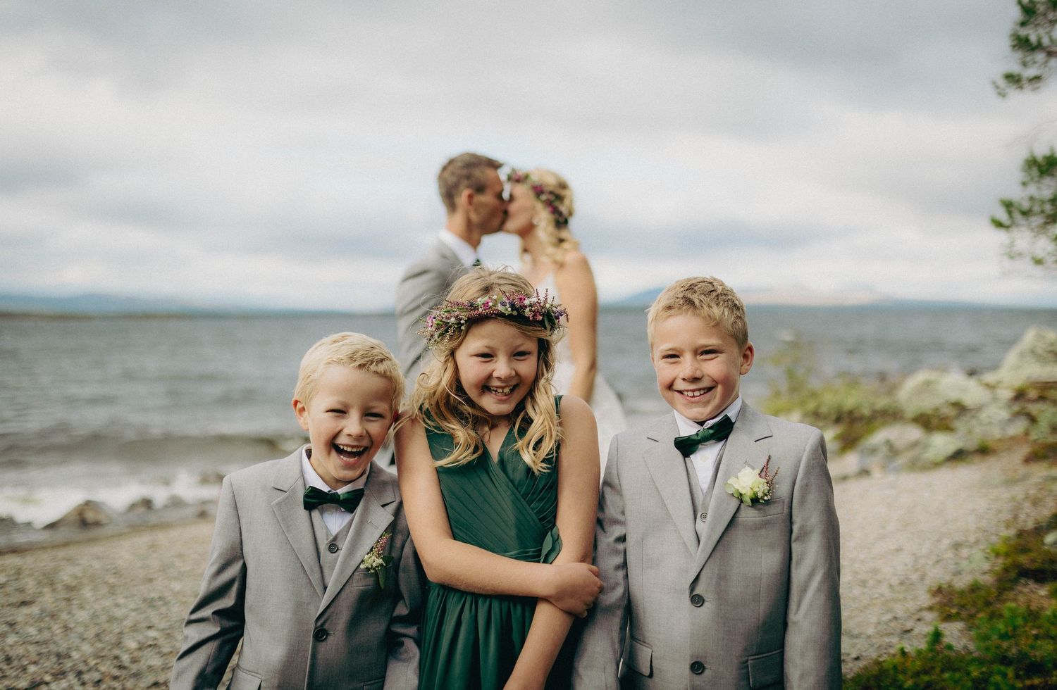 Tre barn som fniser og ler med et brudepar som kysser i bakgrunnen. Morsomt og autentisk familiebilde fotografert under bryllupsdagen.