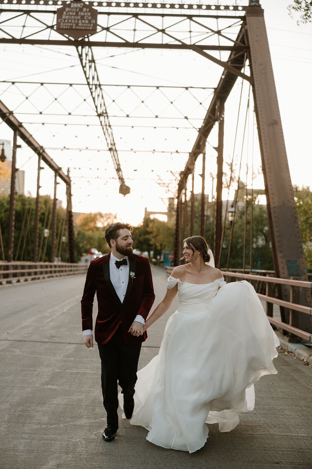 Bride and groom walk hand-in-hand on a historic iron bridge at golden hour, bride in flowing white gown, groom in burgundy tuxedo.