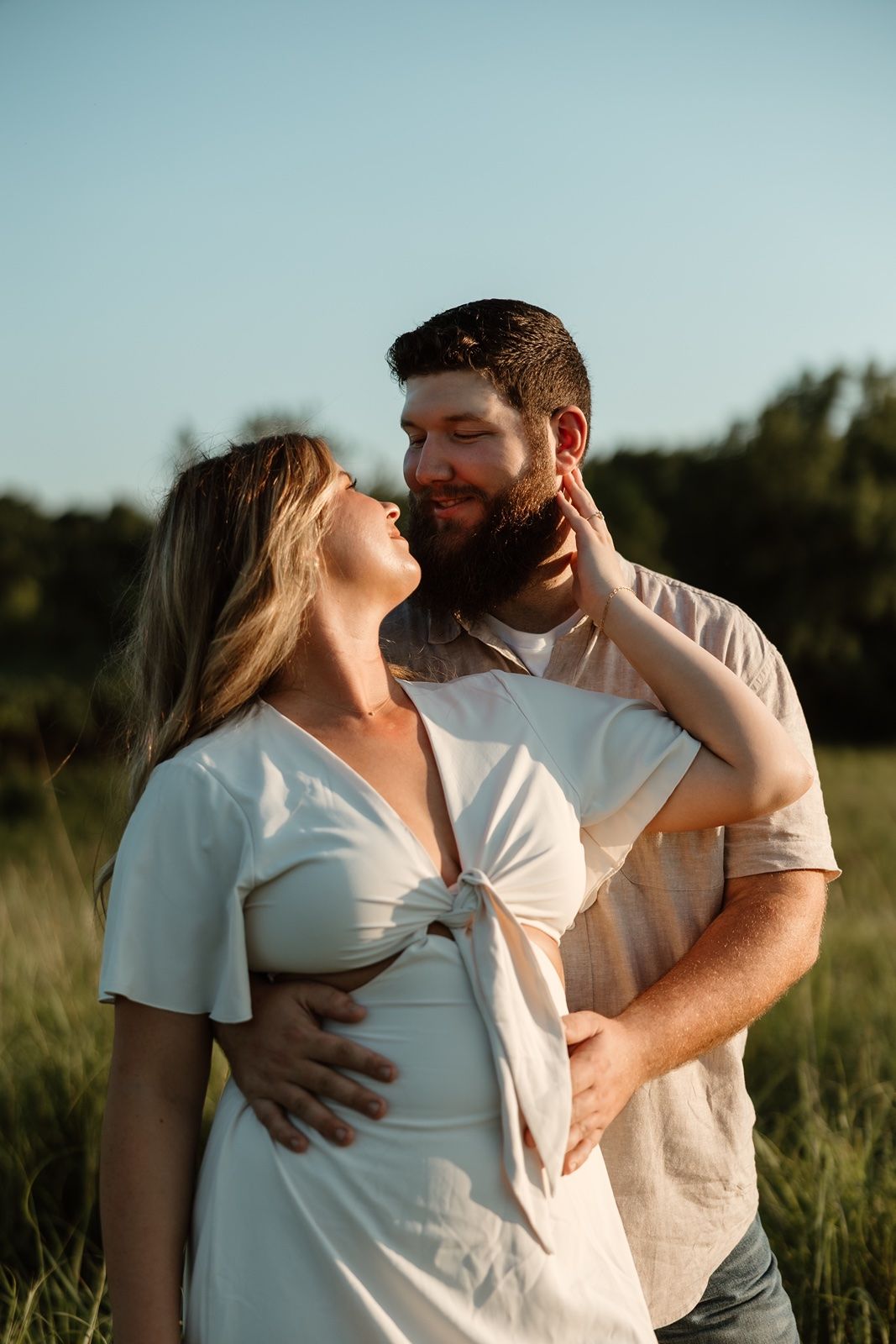 A couple shares a tender moment in a sunlit field, with the woman in a white dress touching the bearded mans face.