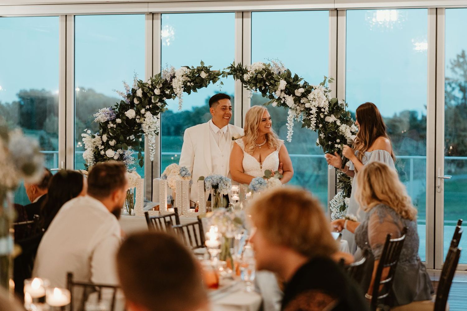 Maid of honor toast at a wedding reception setup with floral arch and guests dining in a sunlit venue with large windows.