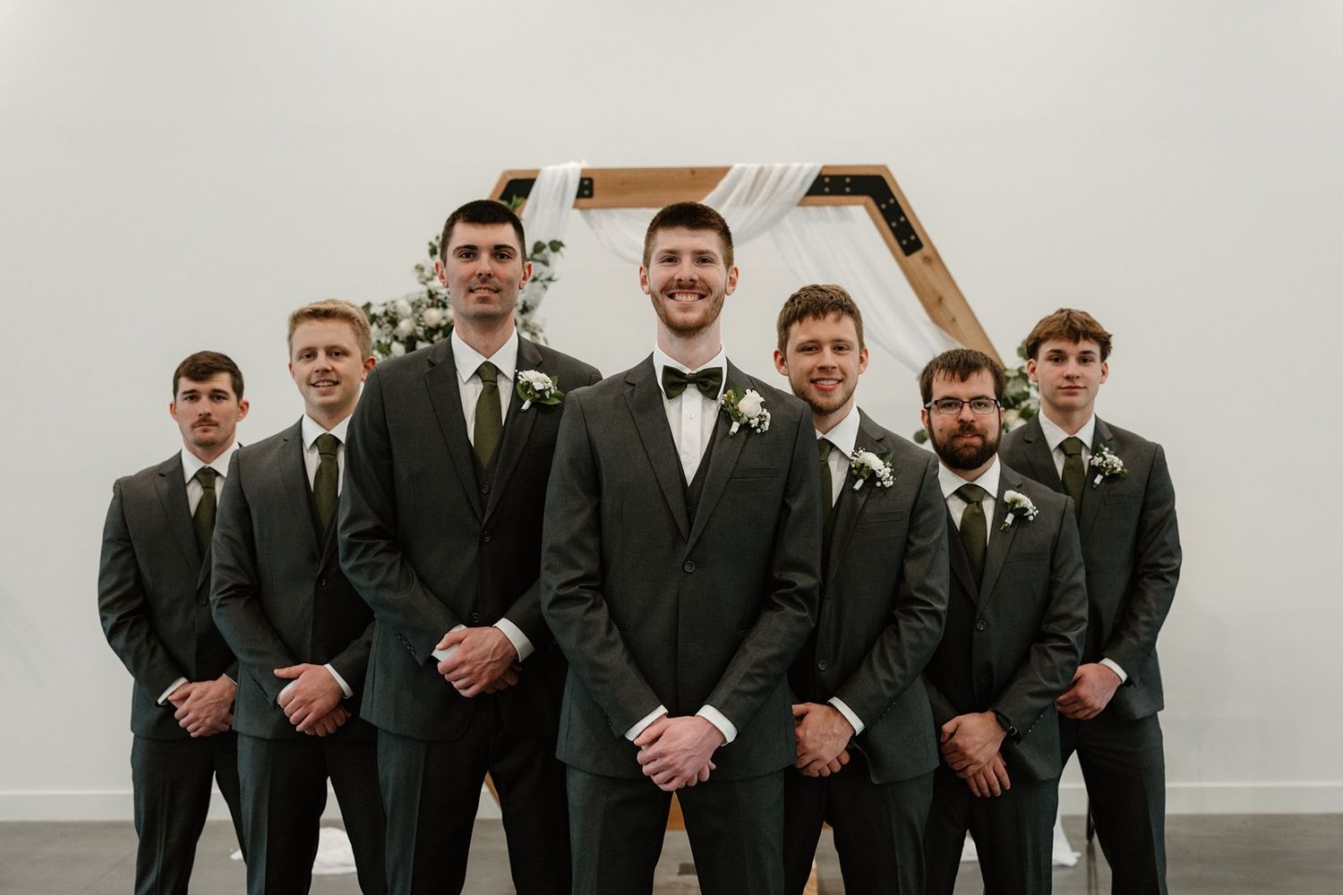 Groomsmen in black suits pose together for a formal wedding photo against a light background.