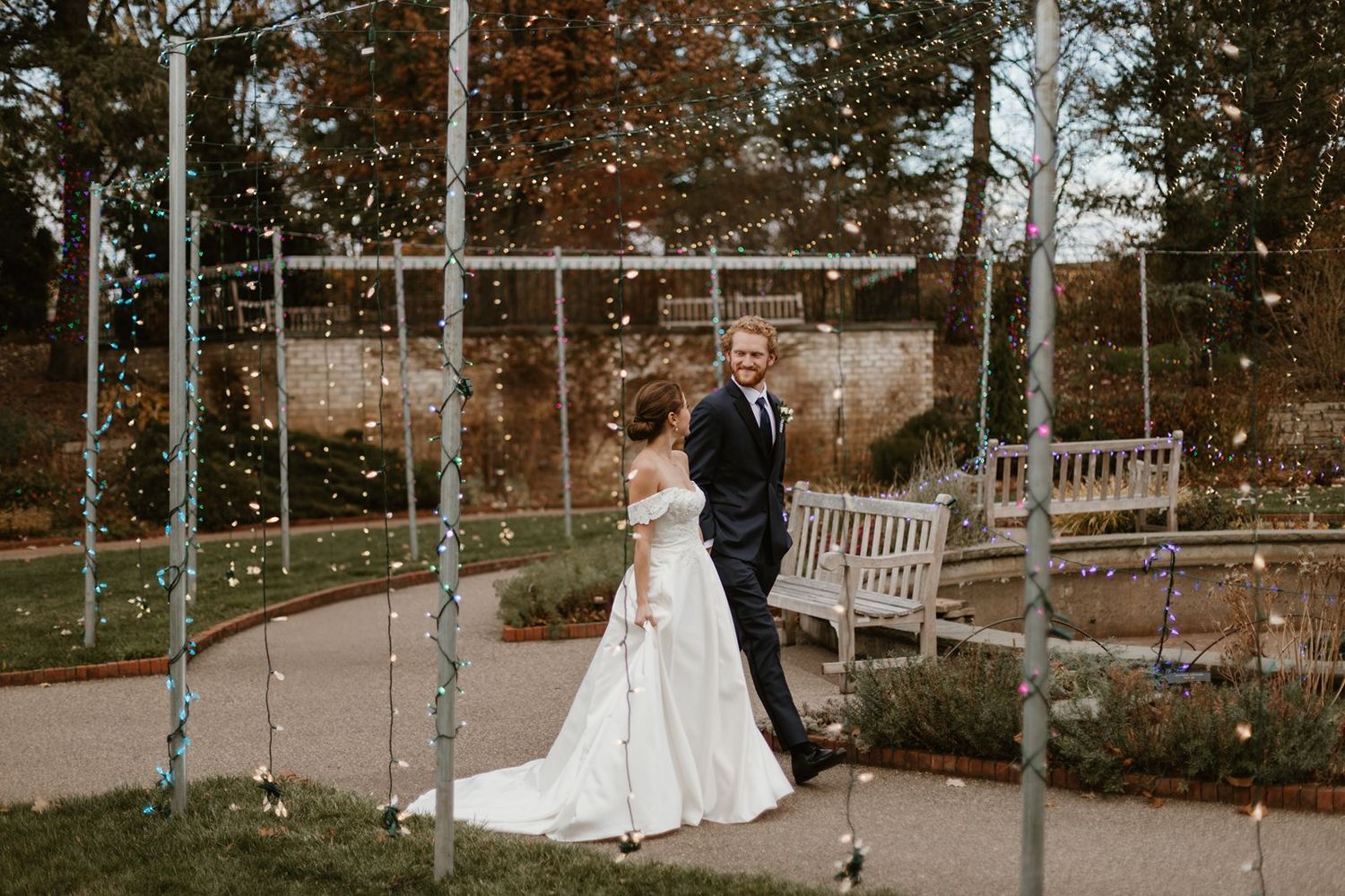 Newlyweds share a sweet moment under string lights and a white lattice structure in an autumn garden setting.