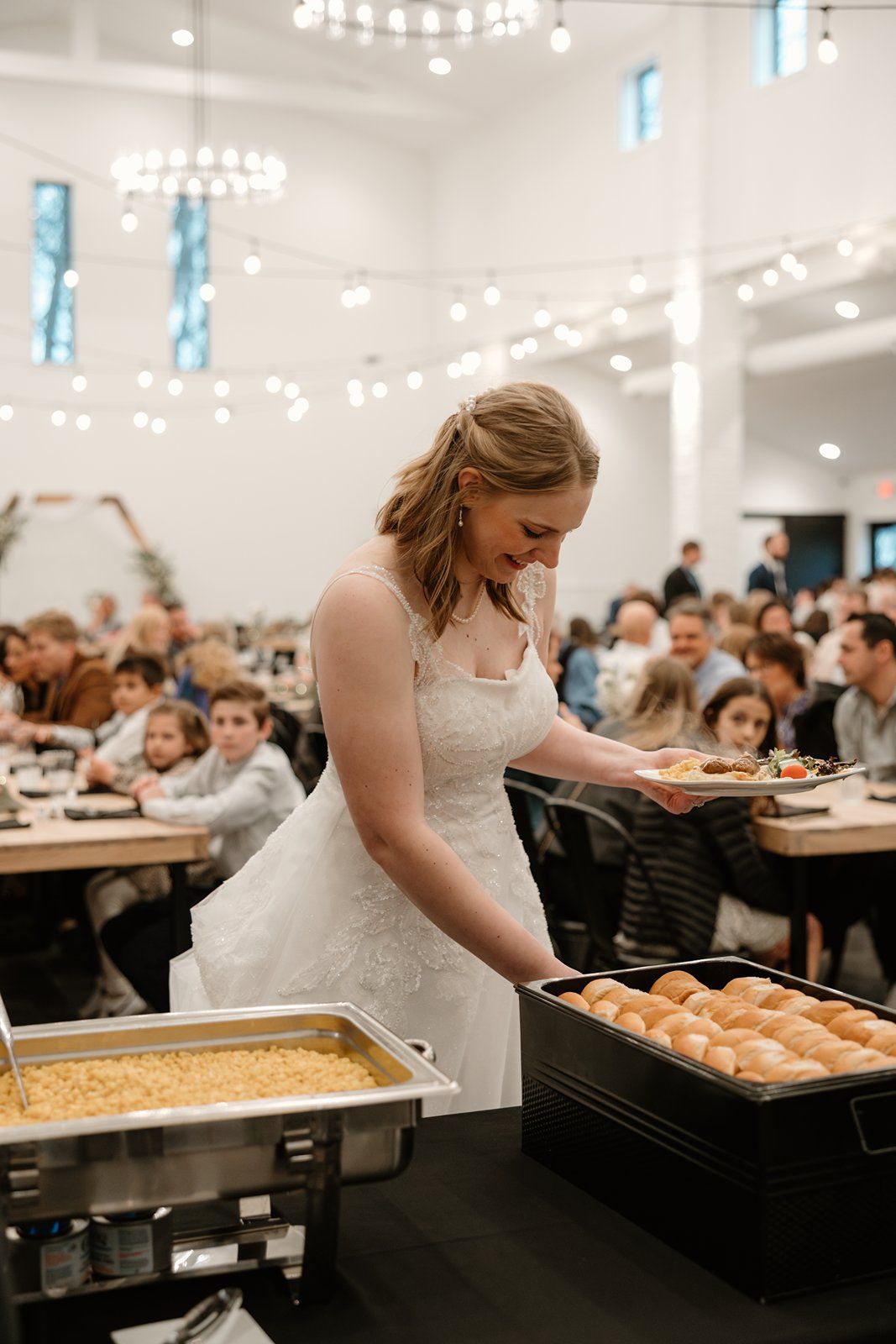 A bride in a white dress serves food from buffet trays at a wedding reception while guests wait in line.