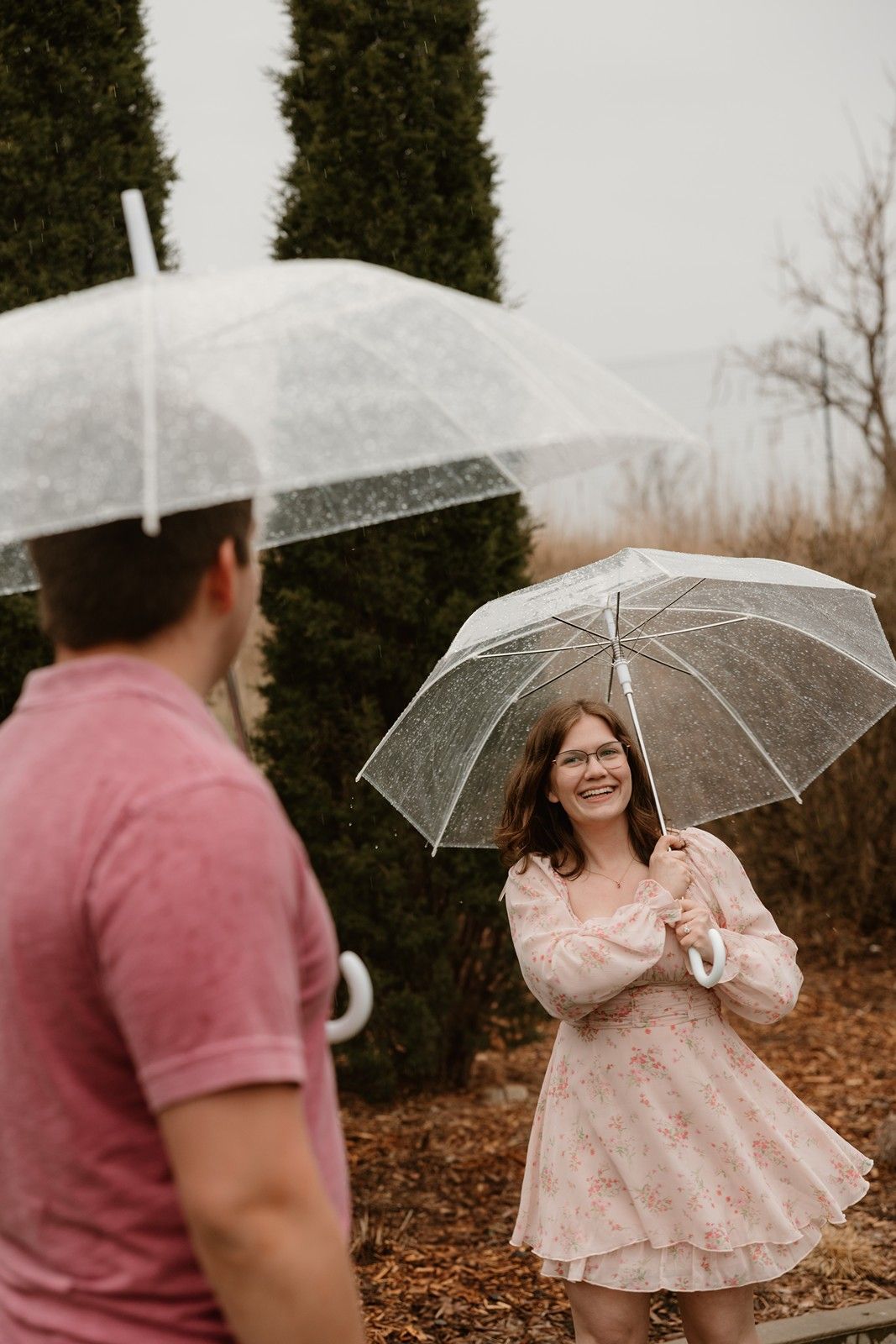Two people holding clear umbrellas stand facing each other on fallen leaves wearing pink colored clothing.