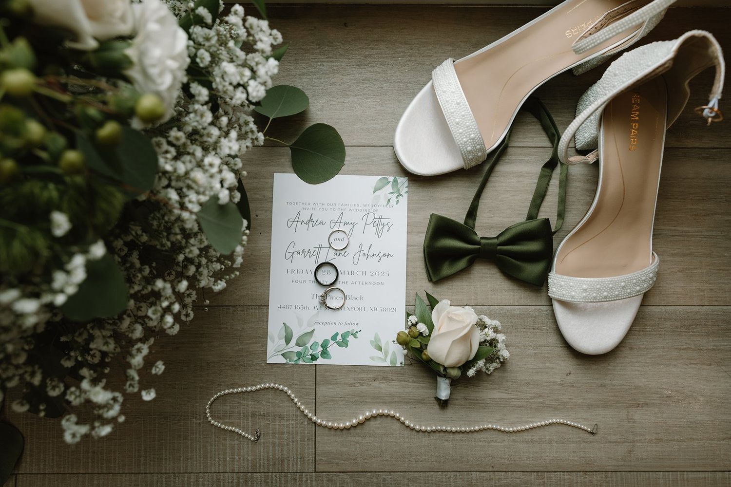 Wedding details flatlay with white invitation card, bridal shoes, rings, and delicate baby's breath flowers on wooden surface.