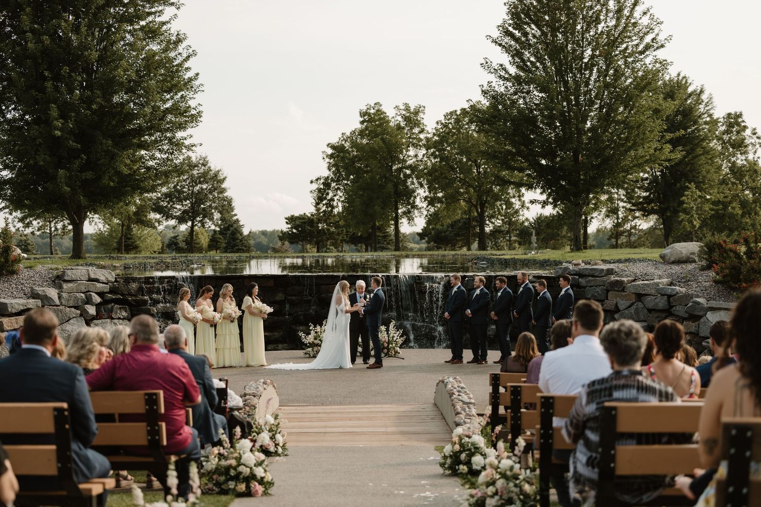 An outdoor wedding ceremony takes place on a stone patio surrounded by trees and wooden benches filled with guests.