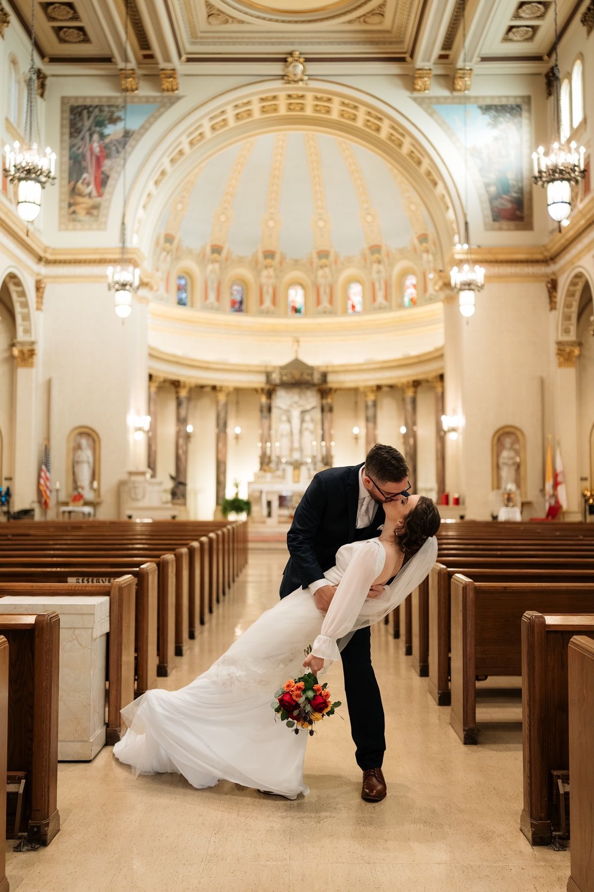 Bride and groom share a romantic dip kiss in the aisle of an ornate church with golden domed ceiling.