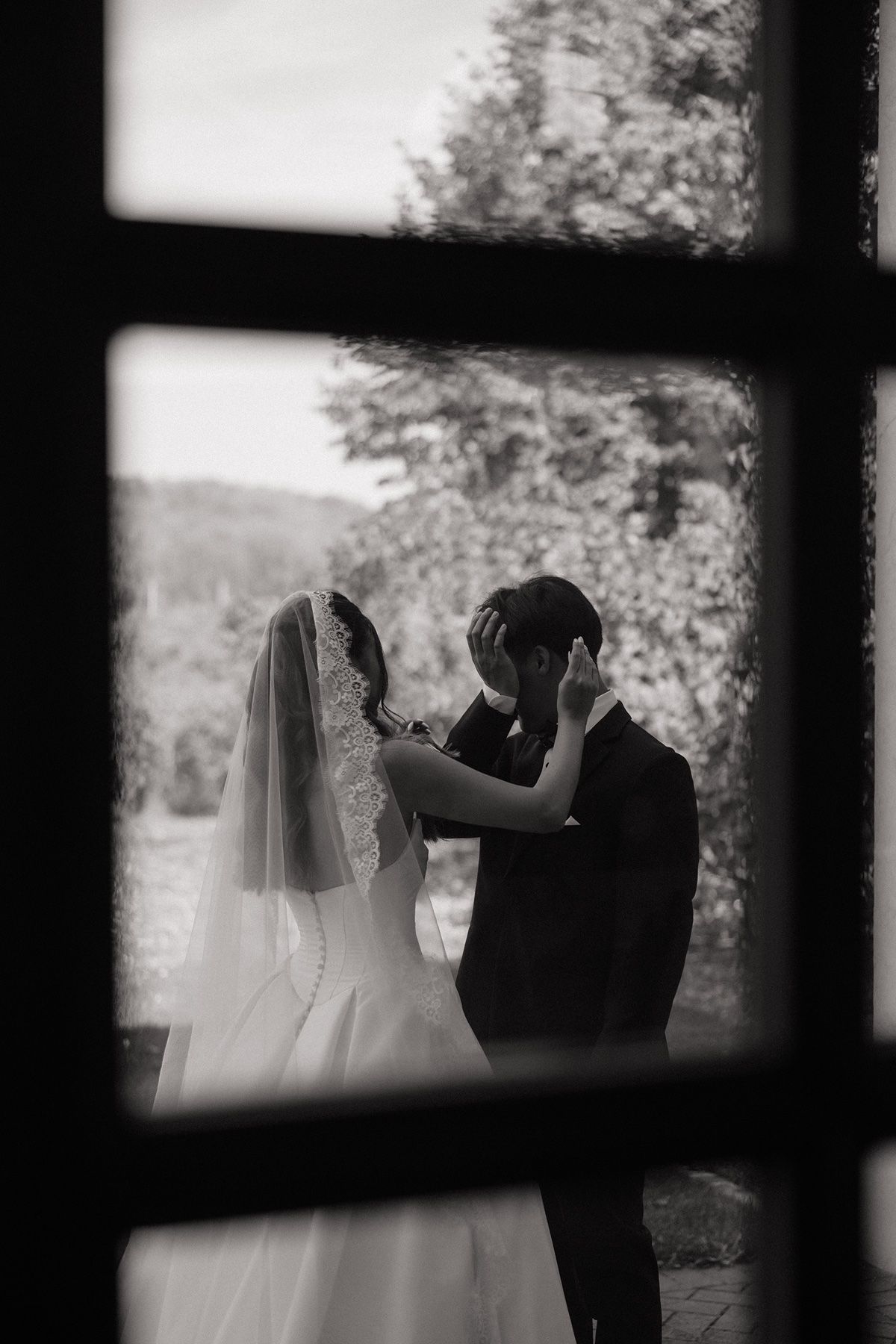 Black and white photo of bride and groom sharing tender moment, viewed through window panes, with lush trees in background.