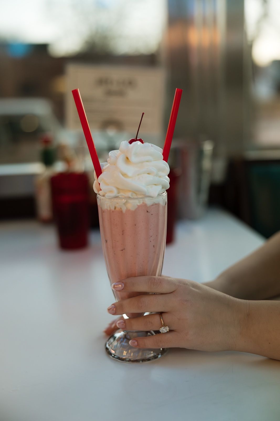 Ring detail pic of a woman holding a pink milkshake topped with whipped cream and a cherry, with two red straws, on a white diner counter.