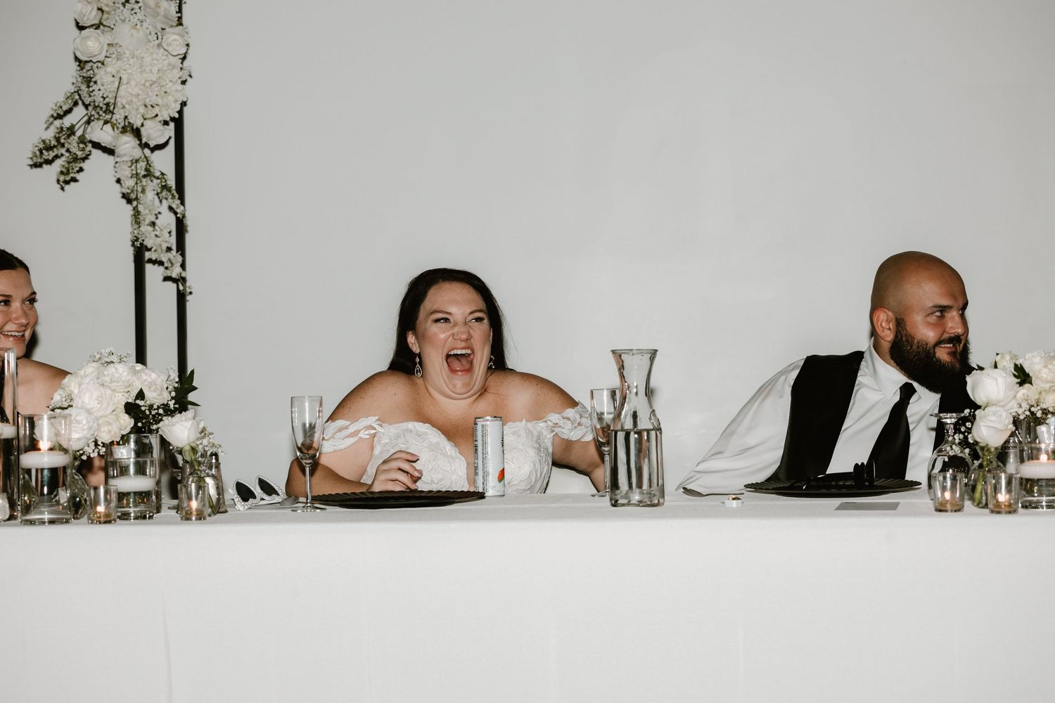 Wedding party sits at head table with bride appearing surprised while someone gives a speech during reception.