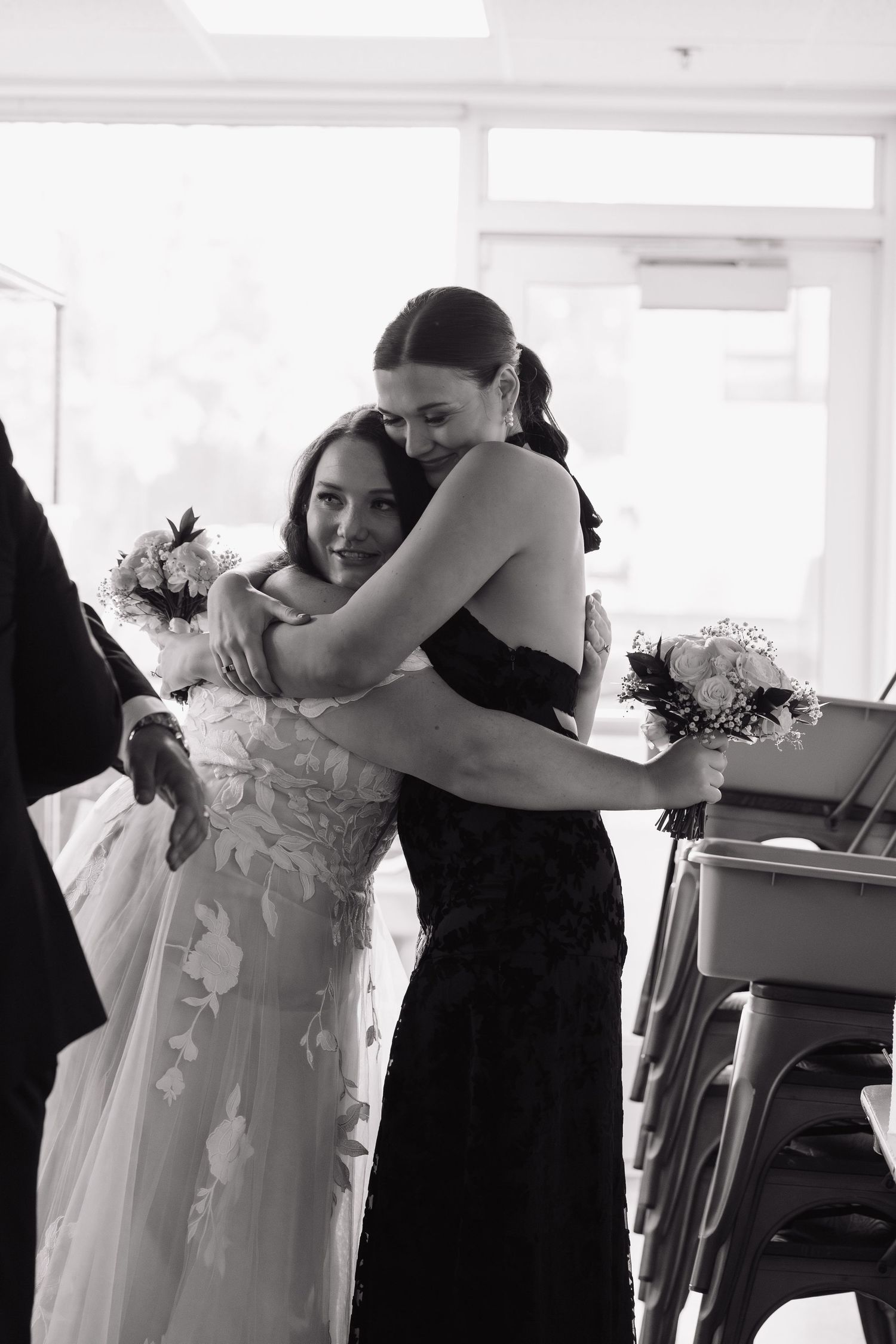 Two women in white and black dresses share an emotional embrace during a wedding celebration.