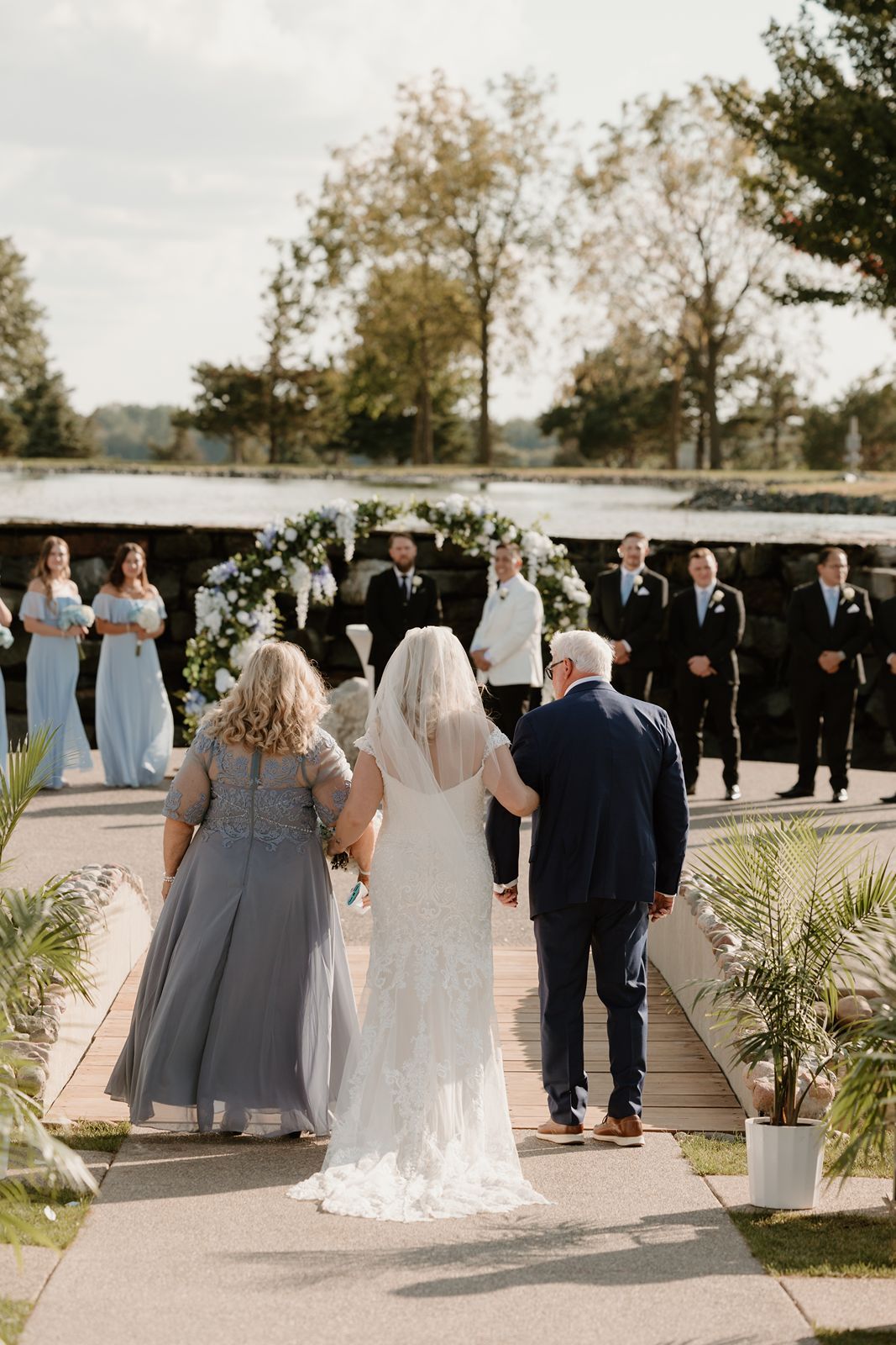 A bride walks down outdoor aisle with wedding party lined up in pastel blue dresses.
