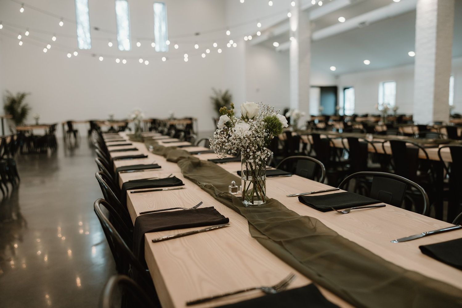 Long banquet tables with white linens and greenery centerpieces set up in a modern venue with string lights overhead.