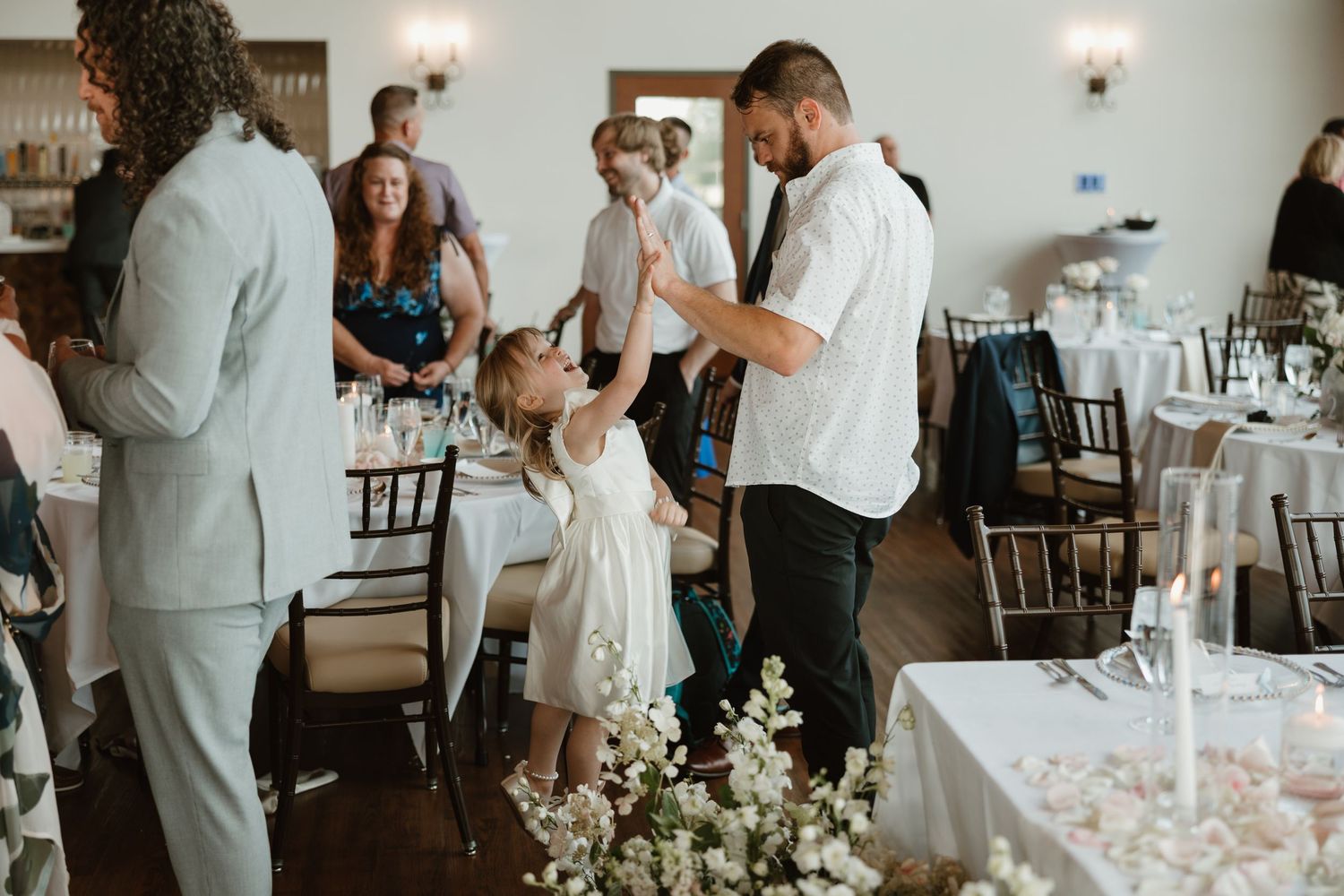 Wedding reception guests mingle and dance in an elegant venue decorated with white floral centerpieces.