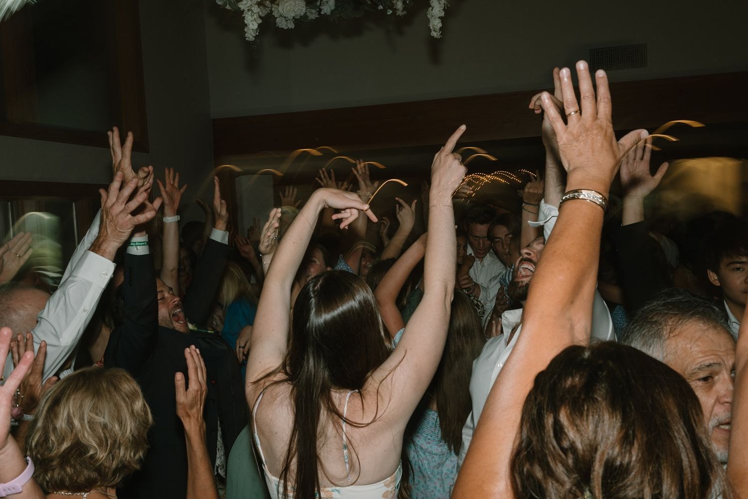Wedding reception guests dancing with hands raised on a dimly lit dance floor during a celebration.