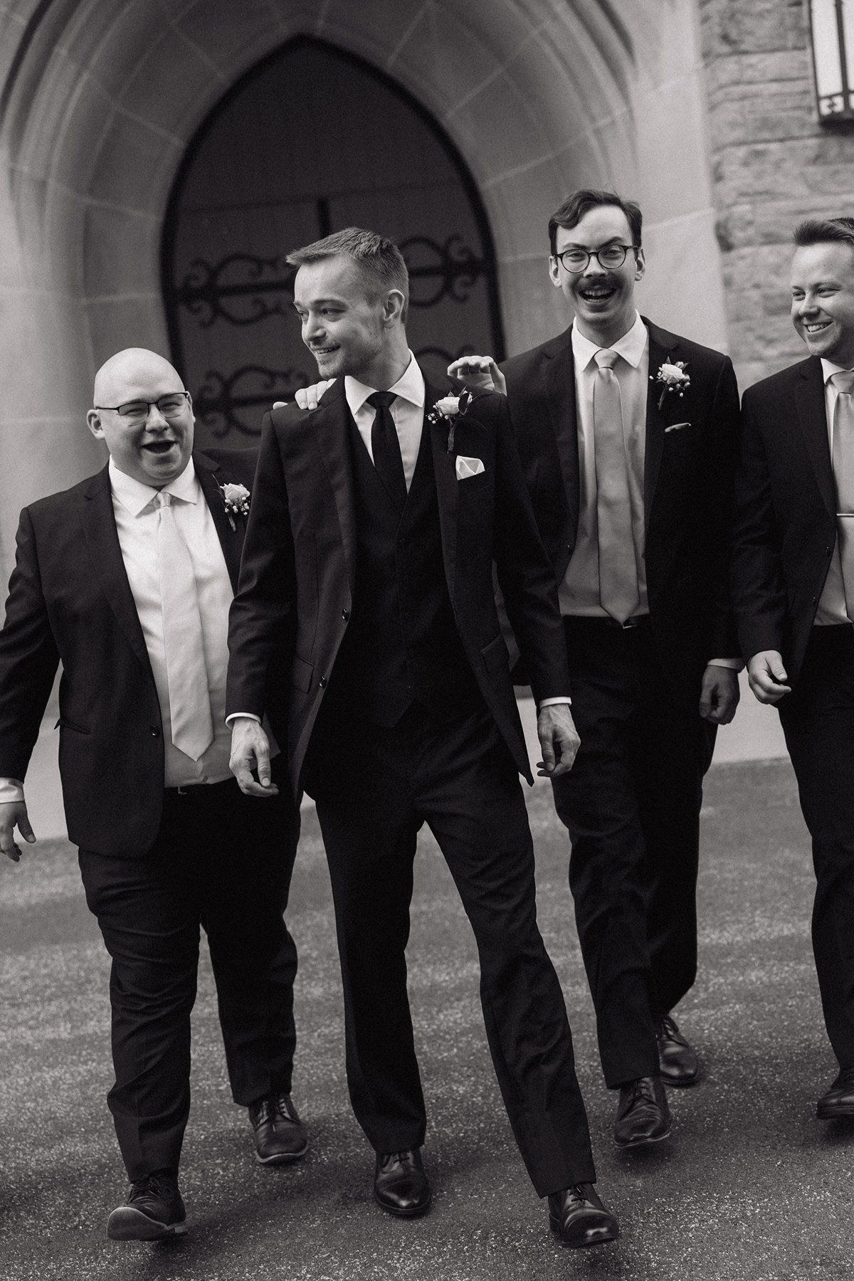 A black and white photo of groomsmen walking and laughing together outside a church entrance.