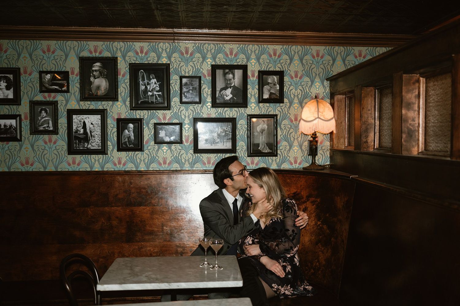 A couple shares an intimate moment in a dark vintage bar with antique photos on teal wallpapered walls and mood lighting.