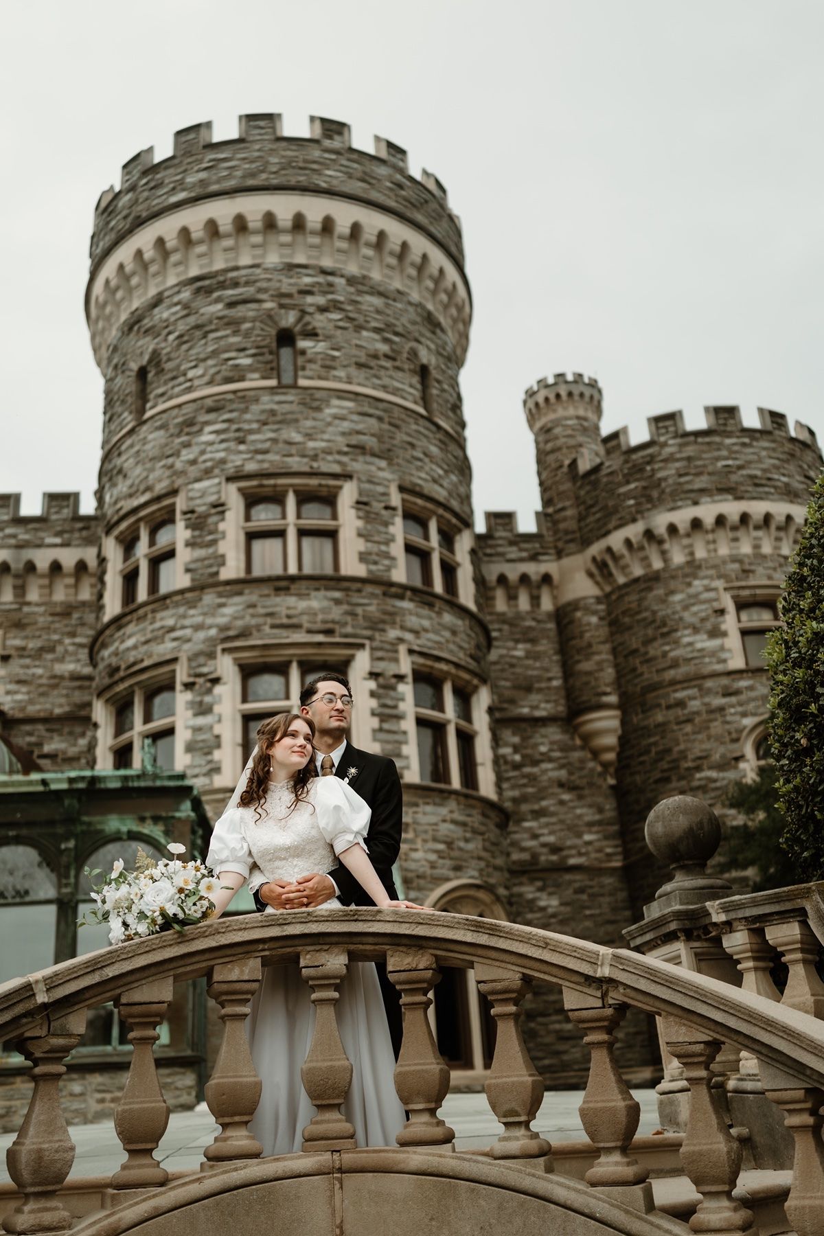 Bride and groom pose on stone staircase before a grand castle backdrop on their wedding day.