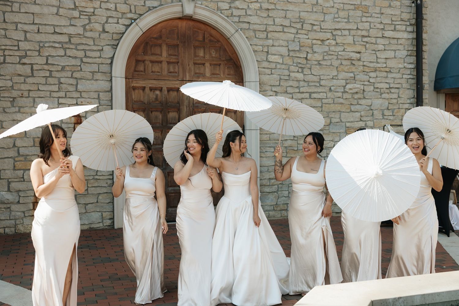 Bridesmaids in white gowns holding parasol umbrellas outside a stone vineyard building.