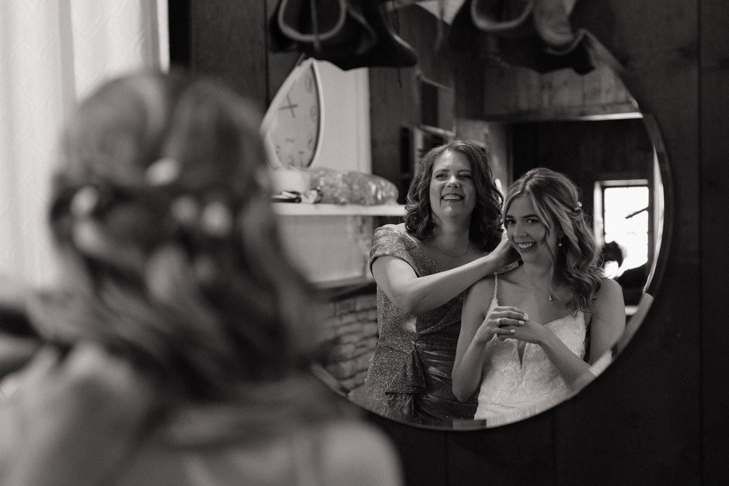 Black and white photo of bride smiling in round mirror reflection while bridesmaid styles her hair before wedding.