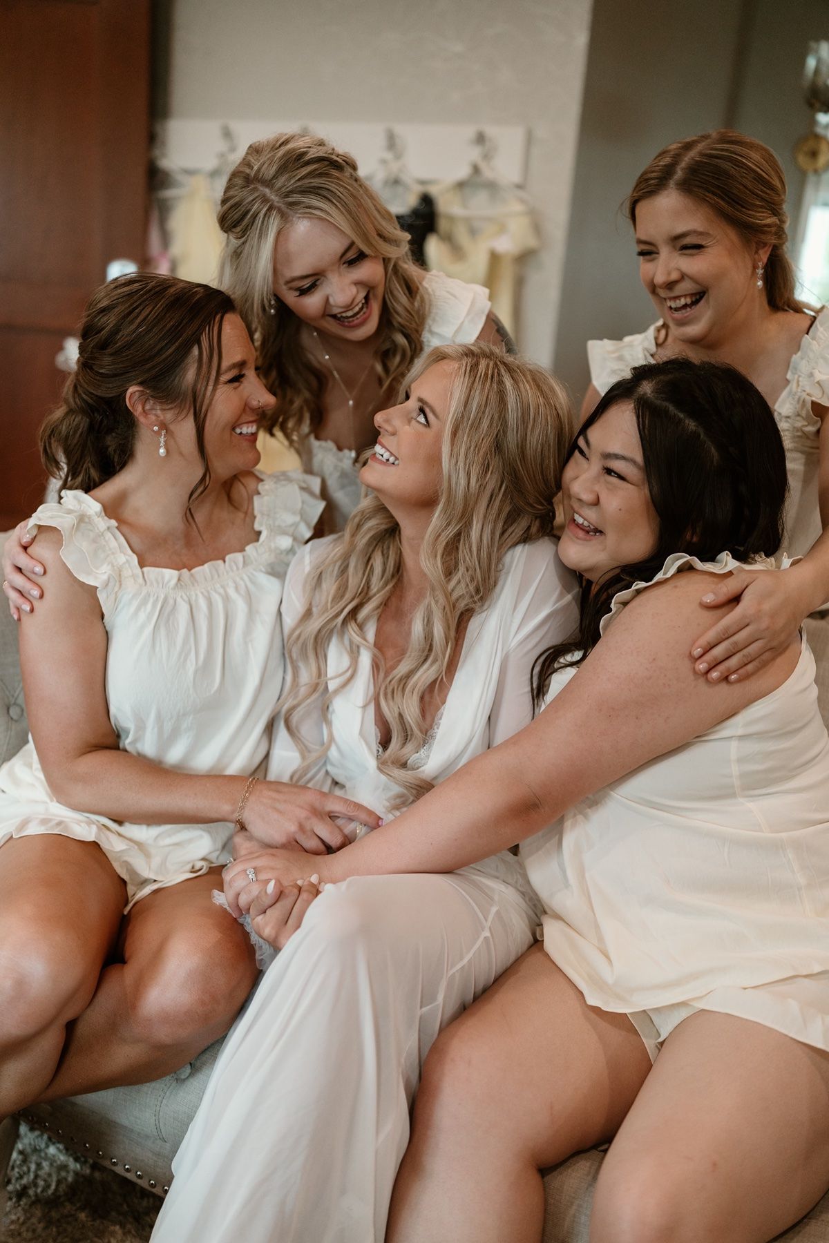 Five bridesmaids in white robes laughing and embracing together during wedding morning preparations.
