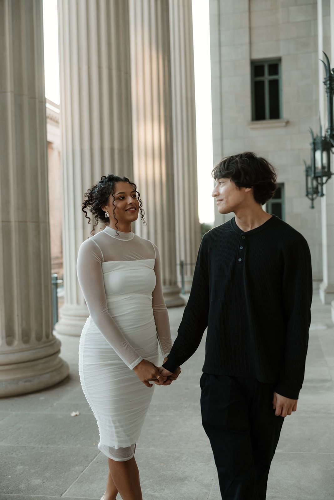 A couple holds hands in front of grand columns, woman in white dress and man in black outfit, smiling at each other.