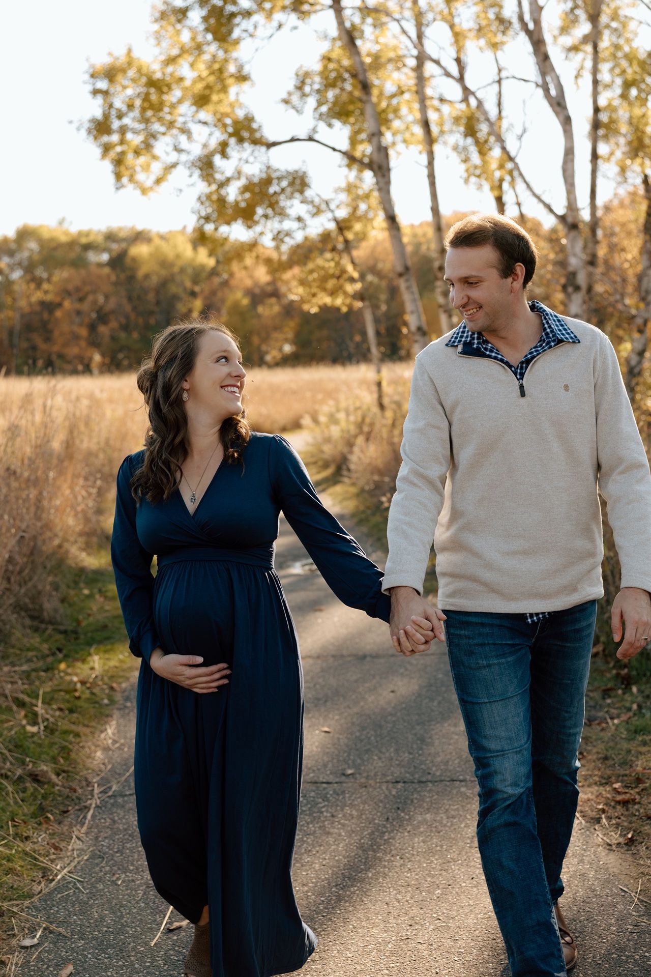 Pregnant couple holding hands and smiling while walking on a path surrounded by autumn trees in golden light.