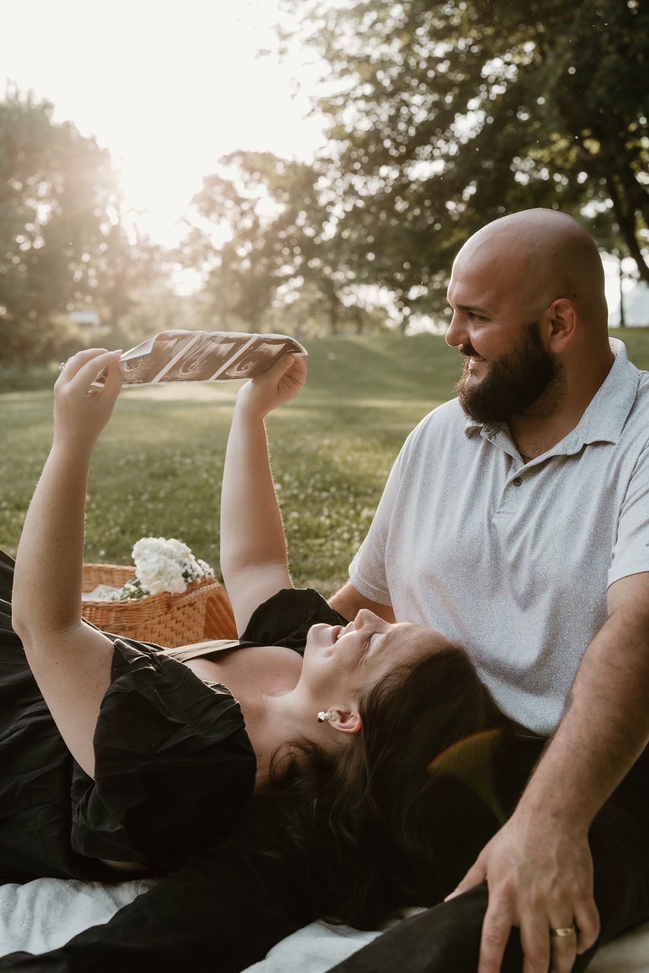Couple relaxing outdoors at golden hour, woman lying down holding ultrasound photos while man smiles warmly beside her.