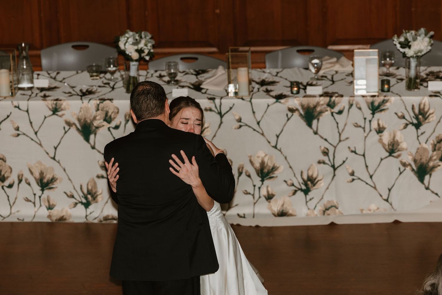 A warm embrace shared during a wedding reception with floral decorations in the background.